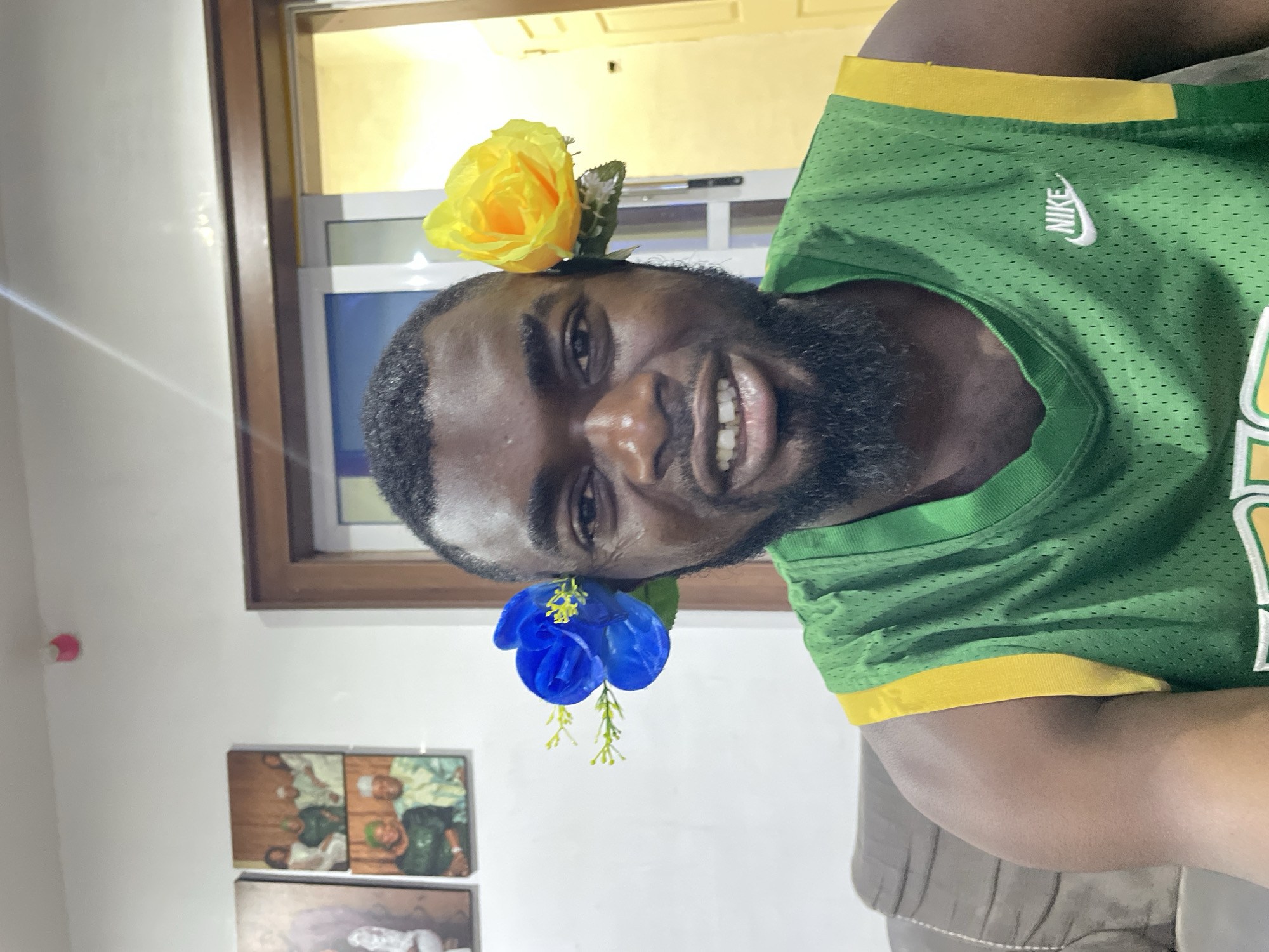 A man is posing indoors, appearing to take a selfie. He has a beard and is wearing a green and yellow Nike athletic shirt. He has a yellow flower placed above his left ear and a blue flower above his right ear. Behind him is a window and two framed pictures on the wall. The location is not explicitly stated as Jos, Nigeria in the image.