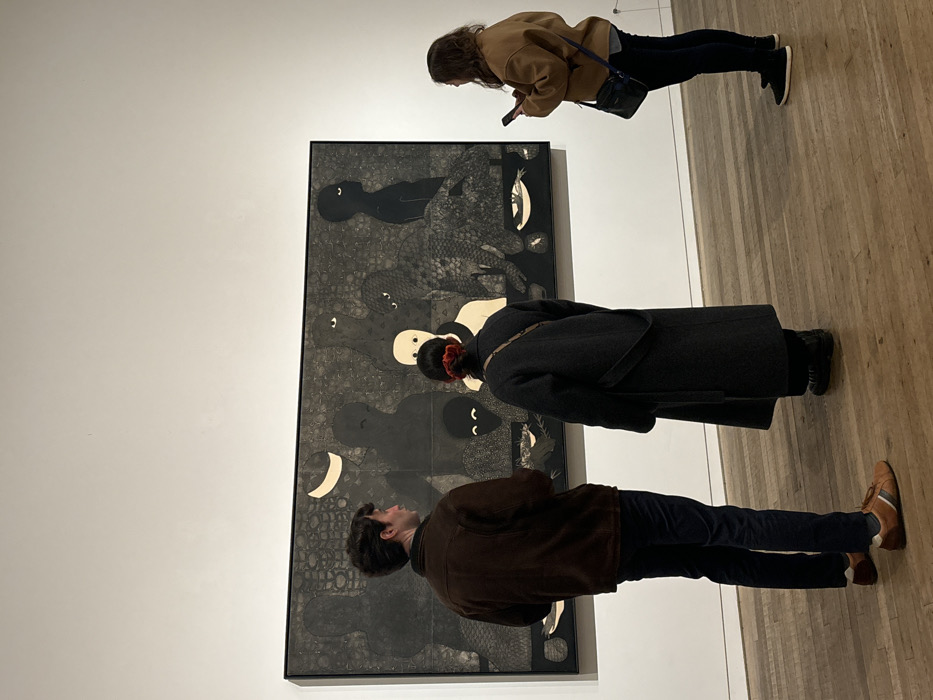 Three individuals are observed in an art gallery at Tate Modern, Bankside, London, United Kingdom. On the left, a male subject in a brown jacket and dark trousers stands facing a large horizontal artwork, looking towards it. In the center, a female subject, her back to the camera, wears a long dark coat and has a red scrunchie in her hair; she is also observing the artwork. On the right, a second female subject in a tan jacket and dark trousers holds a mobile phone with both hands, looking at its screen, with a dark cross-body bag slung over her shoulder. All three stand on a light wooden floor with horizontal planks. The background consists of a plain white gallery wall, upon which the large, framed artwork is mounted. The artwork features a dark, textured background with multiple stylized figures, primarily in black and white, depicting abstract human-like and animal-like forms, including dark silhouettes with eyes, a pale central figure, a crescent moon, and what appear to be fish or birds.