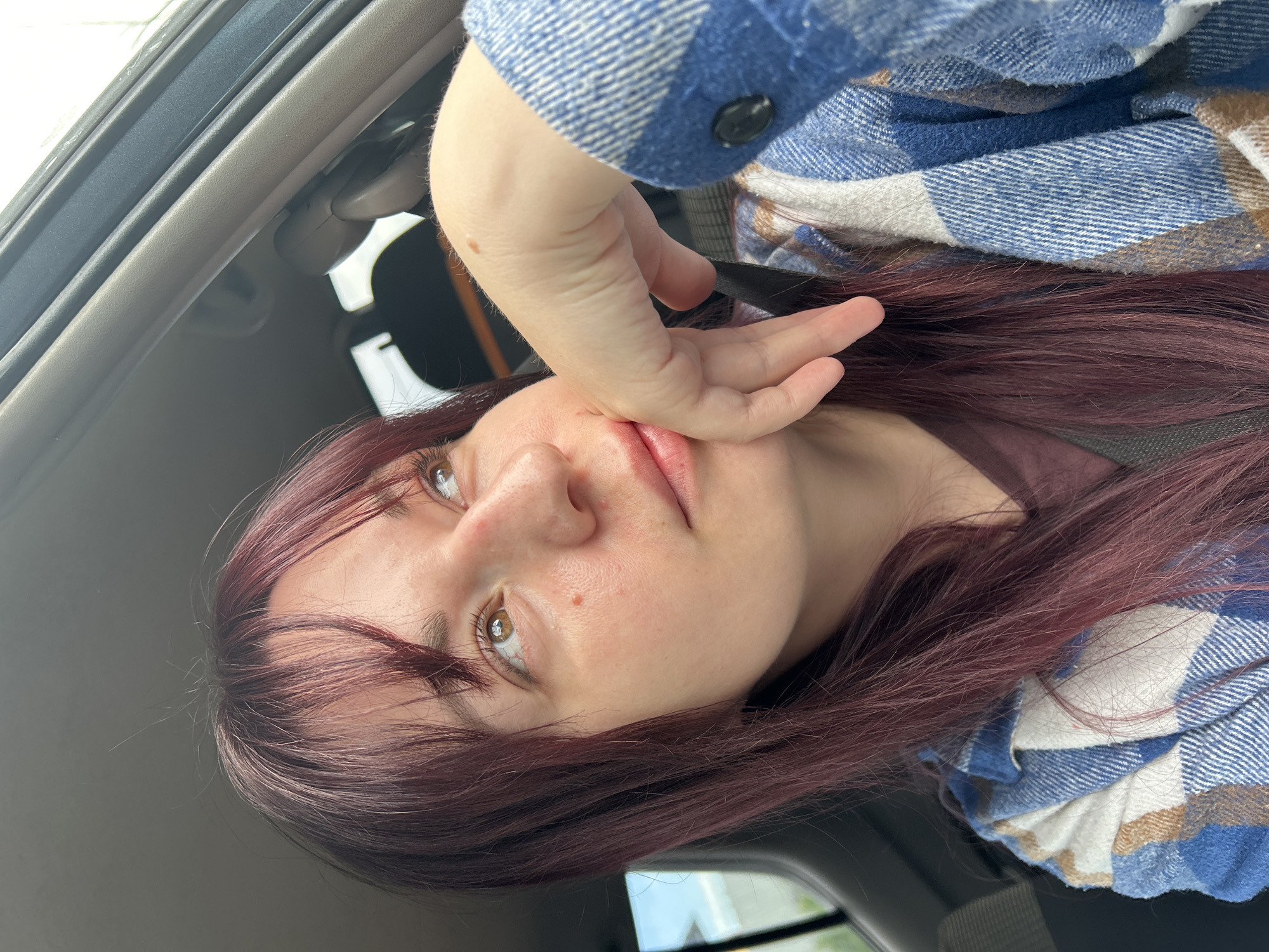 The image shows a close-up, selfie-style shot of a young woman with long, dark red hair and fair skin, looking upwards and to her right. She is inside a vehicle, as indicated by the car interior visible in the background, including the car door frame and a seatbelt. Her left hand is raised, with her fingers gently resting on her chin and cheek. She is wearing a blue and white plaid flannel shirt over a dark reddish-brown t-shirt. The lighting suggests it is daytime. The scene does not provide any specific information or visual cues to determine a location within the city of Fokino, Russia.