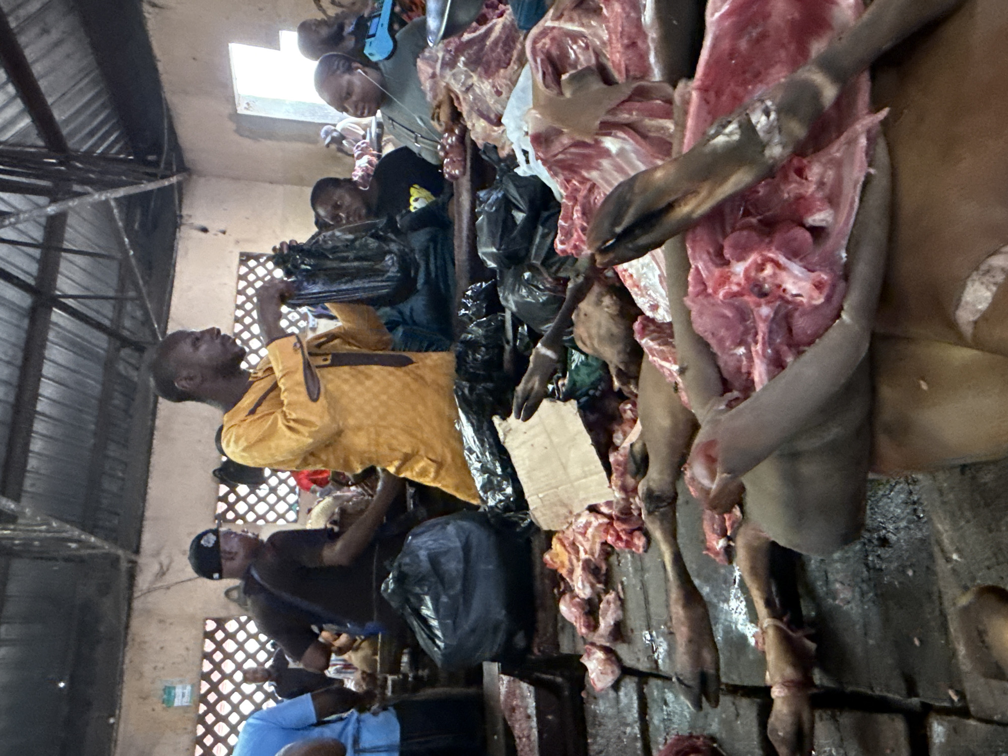 The image depicts a butchery scene in Lugbe, Nigeria. Several individuals are present, primarily engaged in the processing and sale of meat, likely beef or goat. Carcasses or large cuts of meat are visibly displayed and handled. People are seen bagging meat for customers. The environment appears to be a market setting with rudimentary structures and possibly open-air conditions. There are black trash bags and discarded cardboard on the counter. The lighting is somewhat dim, suggesting indoor or covered spaces.