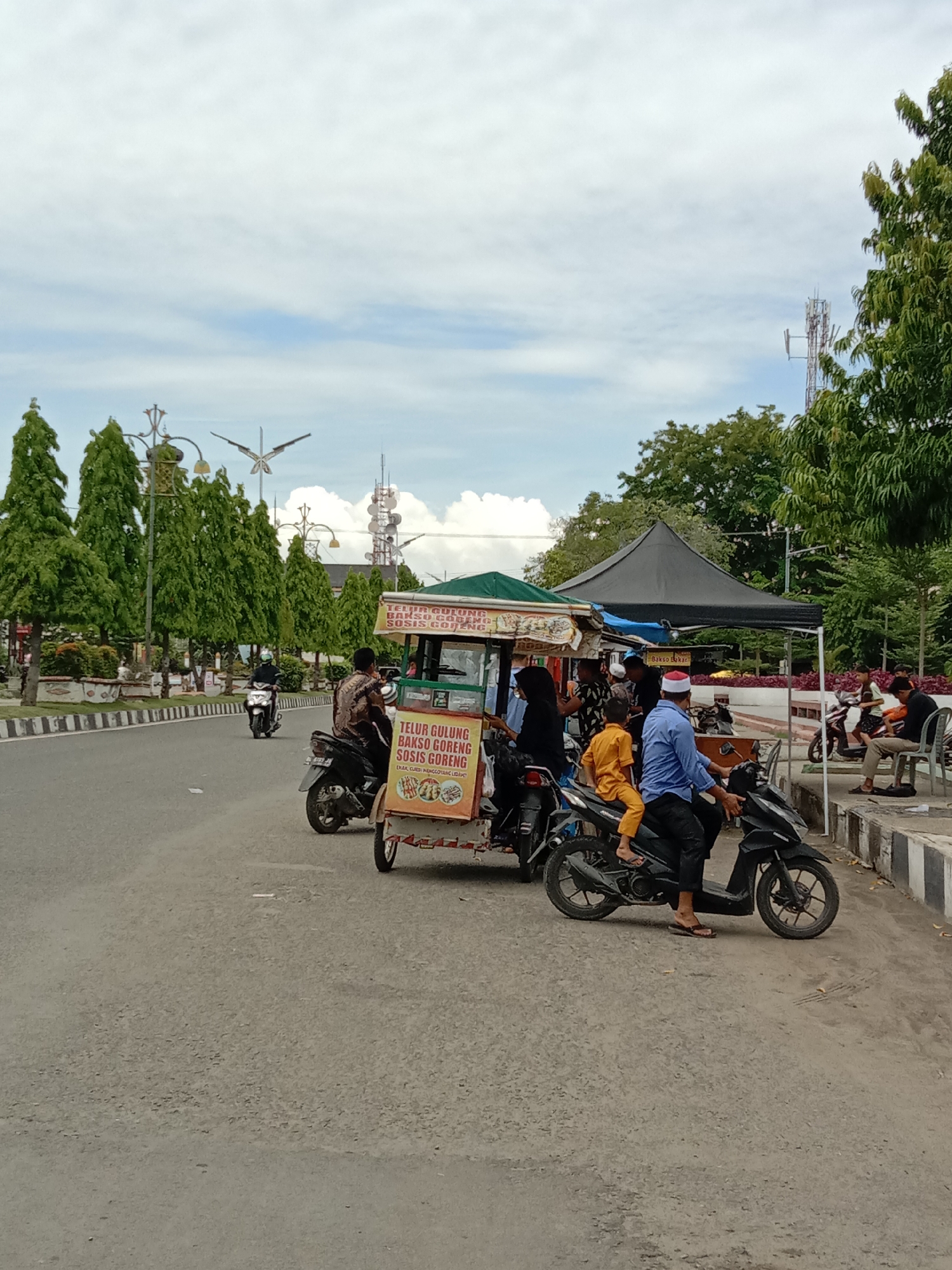 The image shows a street scene in Lhokseumawe, Indonesia, with a food vendor cart parked along the side of the road. The cart advertises "Telur Gulung," "Bakso Cobemo," and "Sosis Goreng" (fried sausage roll, cobemo meatballs, and fried sausage). People on motorbikes are queued up to purchase food from the cart. Nearby, there is a black tent and people are seated in an area with flower beds and trees. Other motorbikes are parked along the curb. Across the street are tall trees, ornate lampposts, and a telecommunications tower. The sky is partly cloudy.