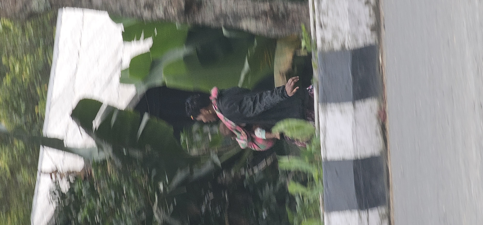 A person is carrying a baby on their back while walking. The person is wearing a dark jacket and a patterned scarf around their neck. The baby is wrapped in a colorful cloth. They are walking on the side of a road with a black and white striped barrier. In the background, there are trees and foliage, and a white structure. The image is blurred due to motion. The location is identified as Puncak Jaya, Indonesia.