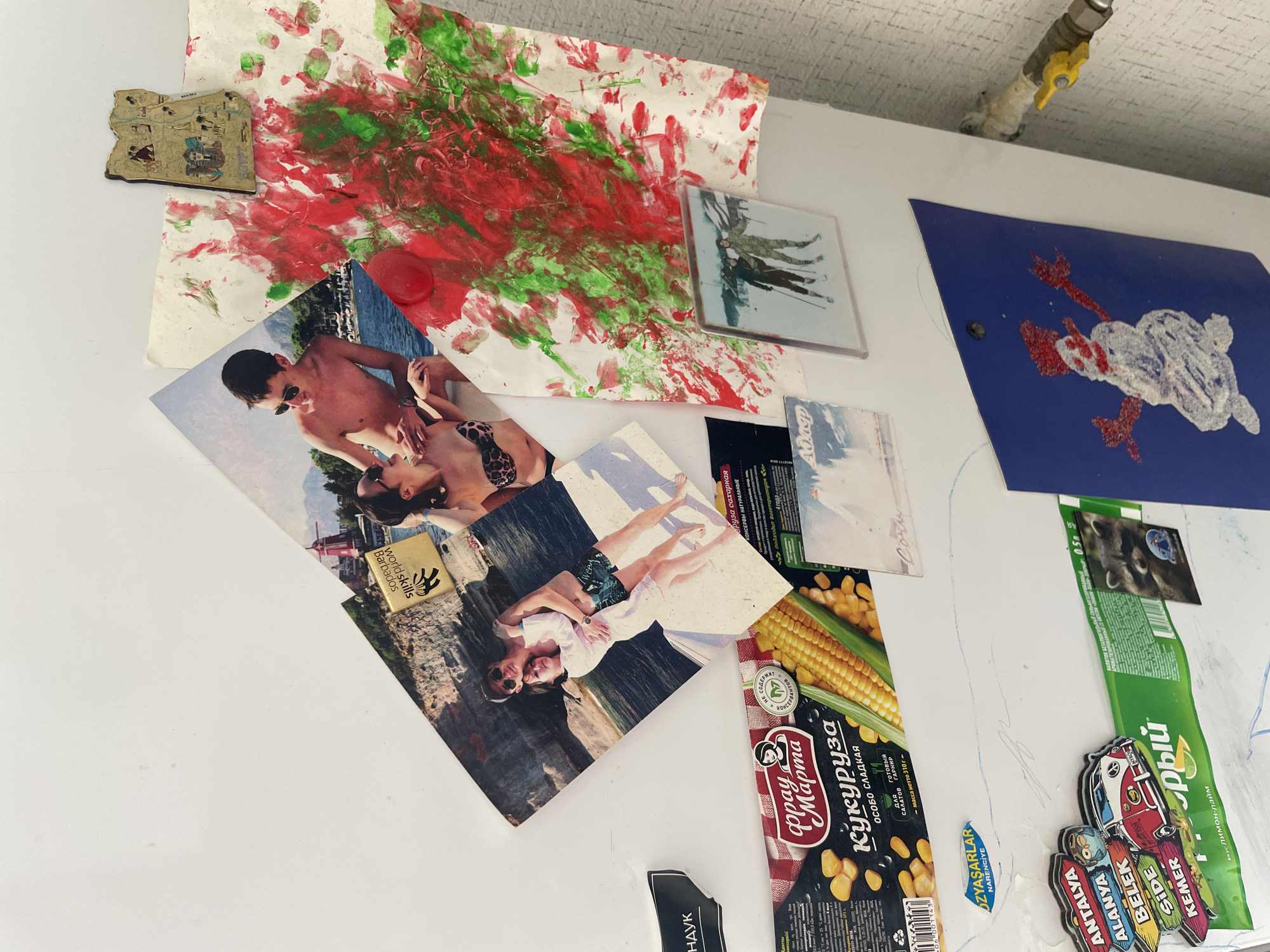 The image shows a close-up of a white refrigerator door adorned with various magnetic items, photographs, and children's artwork. Among the items are several photographs of people, including a couple embracing on a boat and two individuals skiing. There is also a piece of artwork with red and green paint splatters, and a drawing of a snowman on a blue background. Several magnets are visible, including a colorful magnet depicting a van and road signs pointing to different locations in Turkey, as well as a magnet shaped like the country of Egypt. Additionally, there are product packaging for canned corn and a green and white package with text in Russian. A small, framed picture of a raccoon and a snowman is also present. A water pipe with a yellow valve is visible on the right side of the image, emerging from the wall. The background appears to be a textured wall. The image is taken from a slightly elevated angle, looking down onto the refrigerator door. There is no visible indication of the city of Laishevo, Russia within the image itself.