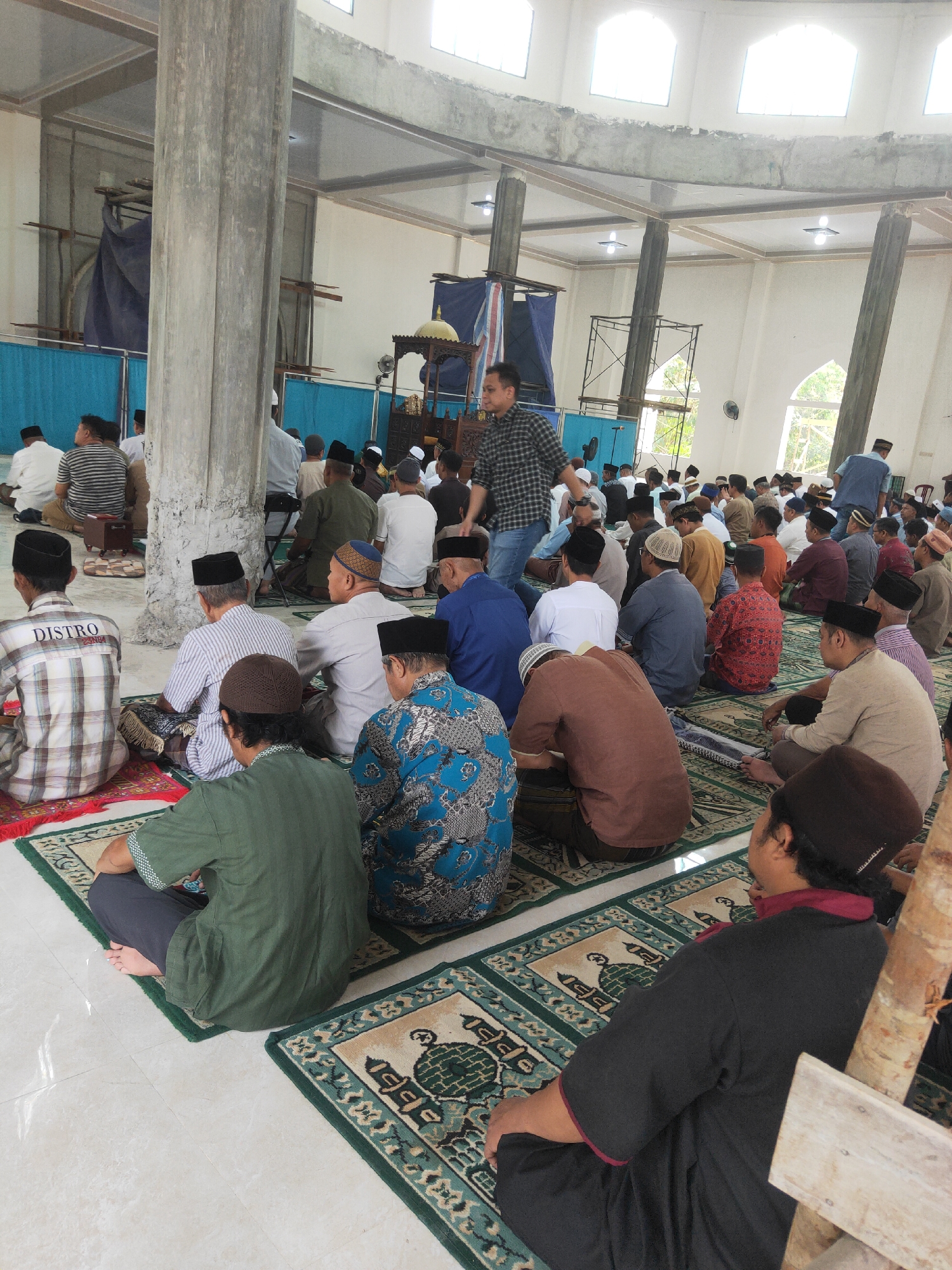 The image depicts an indoor gathering of men, likely for a religious service, taking place in Sintang, Indonesia. The setting appears to be a mosque or a prayer hall, characterized by the presence of prayer mats laid out on a tiled floor. The architecture includes large concrete pillars and arched windows, with scaffolding and construction materials visible in the background, suggesting ongoing renovations or construction.
A significant number of men are seated on the prayer mats, facing in the same direction. They are dressed in various traditional and casual attire, predominantly in muted colors, with many wearing skullcaps or peci. Some men are observed in a seated prayer posture, while others are looking around or interacting with those nearby.
In the foreground, several men are prominently featured, their backs turned to the camera. One man in a green shirt and dark pants is seated on a prayer mat. To his right, another man in a black robe with red trim is seated on a mat. Further to the right, a wooden beam and part of a wooden structure are visible.
In the midground, a group of men are seated. One man in a plaid shirt with the word "DISTRO" visible on the back is seated on a red and white rug. To his left, a man in a striped shirt is also seated. Behind them, more men are positioned on prayer mats, forming rows.
The background reveals more congregants, with some standing and others seated. A man in a checkered shirt and jeans is standing and walking towards the left side of the frame. To his right, a pulpit or minbar with decorative elements and a golden dome is visible. Blue tarps and scaffolding are present, indicating construction or maintenance work. Natural light enters through the arched windows, illuminating the space. The overall impression is one of a community gathering for prayer or a religious event in a partially completed or under renovation building.
