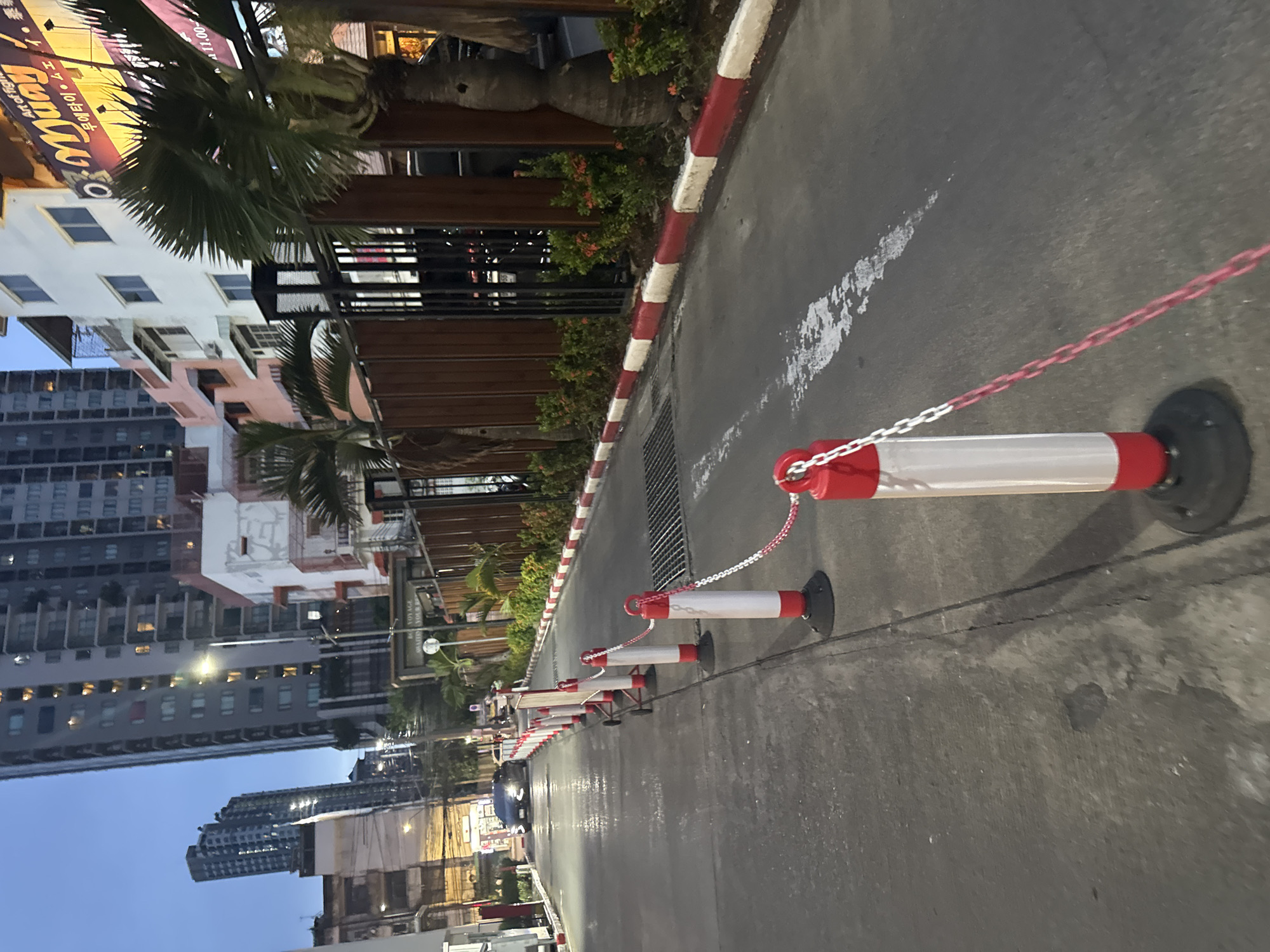 The image depicts a narrow street in Bangkok, Thailand, at dusk or early evening. The street is lined with a series of red and white traffic cones connected by chains, creating a temporary barrier or demarcation. The road surface is asphalt and appears wet, possibly from recent rain. On the left side of the street, there are several multi-story buildings, some with balconies and lit windows, suggesting residential or commercial use. A dark-colored car is visible further down the street, its headlights on. On the right side, a wooden fence runs along the property line, behind which are palm trees and dense green foliage with small red flowers. A sign with Thai script and possibly an advertisement for "Muay Thai" is also visible on the right. The sky is a muted blue, indicating twilight. The scene suggests a typical urban environment in Bangkok, with a mix of modern buildings and tropical vegetation.