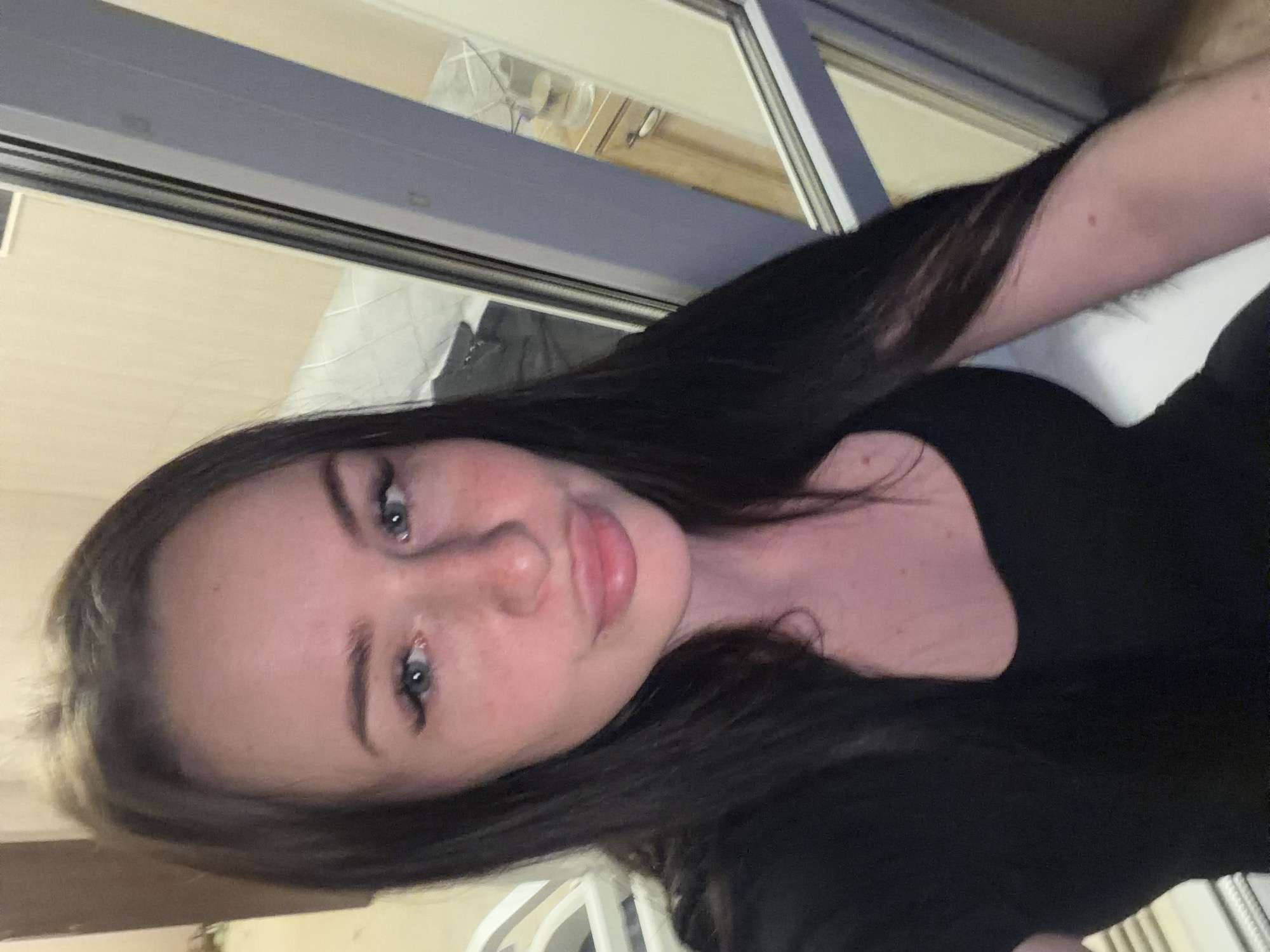 A fair-skinned female with long, dark brown hair and blue eyes is depicted in a headshot-style portrait, looking directly at the camera. Her lips are notably full and pursed in a 'pouting' expression. Small freckles are visible across her nose and cheeks. She wears a black top with a scoop neckline. A small, reddish mark is present on her right arm. The background to her immediate left features a blurred white object and a light-colored wall. To her right, a window with a grey frame reflects an interior space. This reflection shows a light beige wall, a white quilted surface (consistent with a bed or couch) with a dark item resting upon it, and a light wood cabinet with a metal handle. On the cabinet, a clear glass filled with liquid is visible. The scene is illuminated by ambient indoor lighting. There is no information within the image to identify the specific location as Saint Petersburg, Russia.