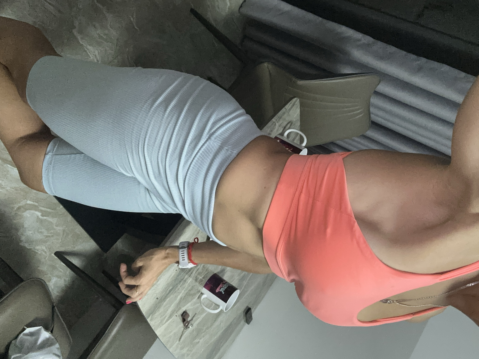 A woman is shown from the waist up in the image. She is wearing a peach-colored workout top and gray compression shorts. A coffee mug, keys, and a digital watch are on a table next to her. Part of a chair and a window with blinds are visible in the background. The location is indicated as Bac Nha Trang Ward, Vietnam.