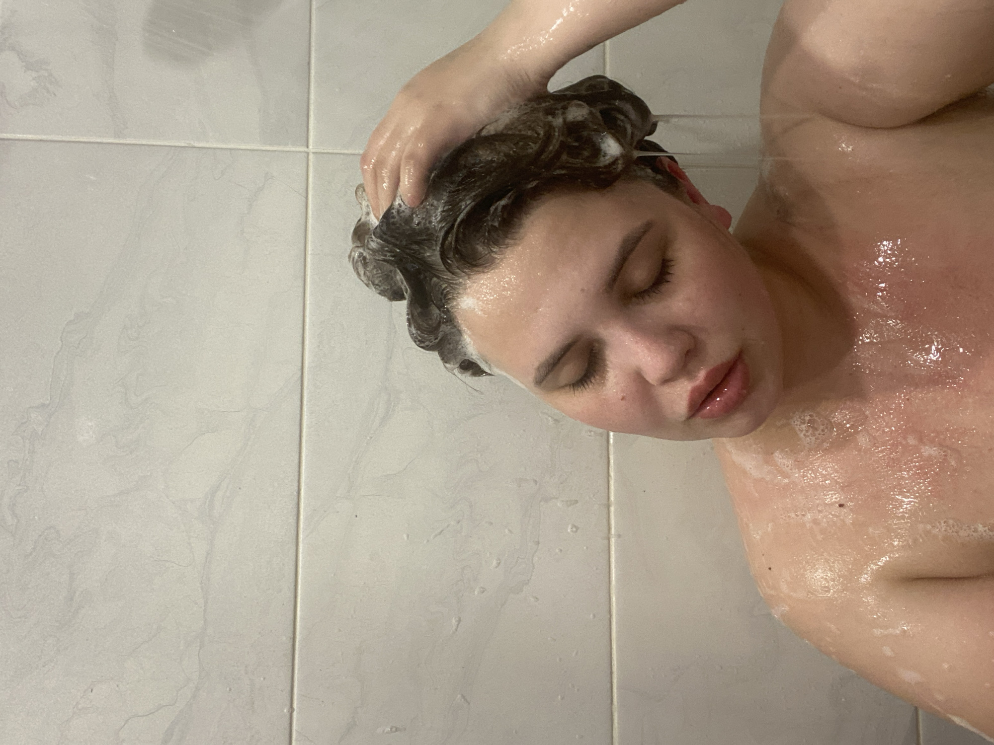An overhead or high-angle shot captures a person washing their hair in a shower or bathroom, located in Chernushka, Russia. The individual, seen from the head to the upper torso, has dark, curly hair thoroughly lathered with white shampoo or soap. One hand is visible, massaging the scalp and hair, creating foam. The person's eyes are closed, and their lips are slightly parted, suggesting a moment of relaxation or concentration during the wash. Their skin, particularly on the shoulder and upper chest, is wet and covered with water droplets and soap suds. The skin appears somewhat flushed.
The background consists of light-colored, rectangular tiles with visible grout lines, typical of a bathroom or shower stall. The tiles have a subtle, light grey marbled or swirled pattern. The lighting appears to be indoor artificial light. No specific time of day or weather conditions are discernible, and there is no visible text in the image. The overall scene conveys a private moment of personal hygiene.