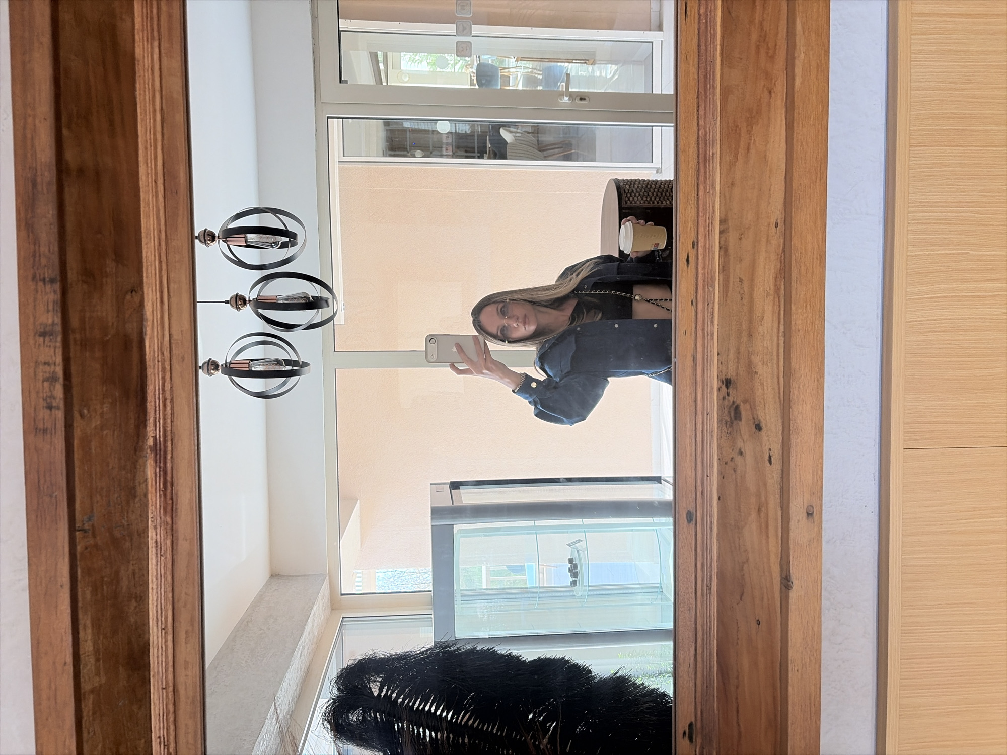 A mirror reflects a person holding a phone and a cup, a light fixture, a display case, and a view through a window. The location appears to be an interior space with modern design elements.