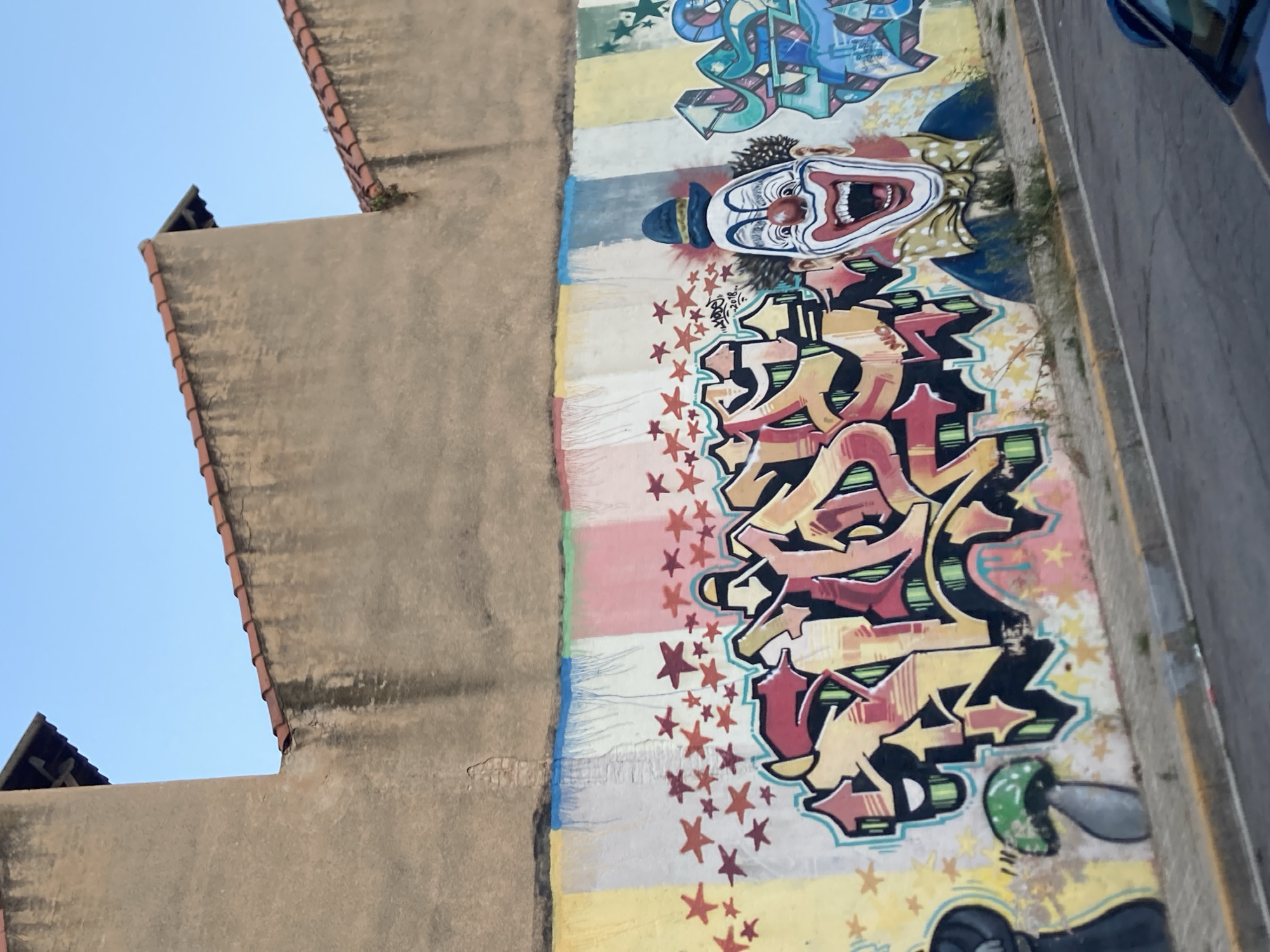 This is a photograph of a graffiti-covered wall in Badalona, Spain. The wall features a colorful, abstract mural with various elements including a large, cartoonish clown with a wide-open mouth and a blue hat, as well as large, stylized lettering in red and yellow. Scattered across the wall are numerous small red and yellow stars. The wall itself has a striped pattern of pastel colors at the top, just below a tan-colored, textured building with a red-tiled roof. The bottom of the image shows a sidewalk and a street with a car partially visible.