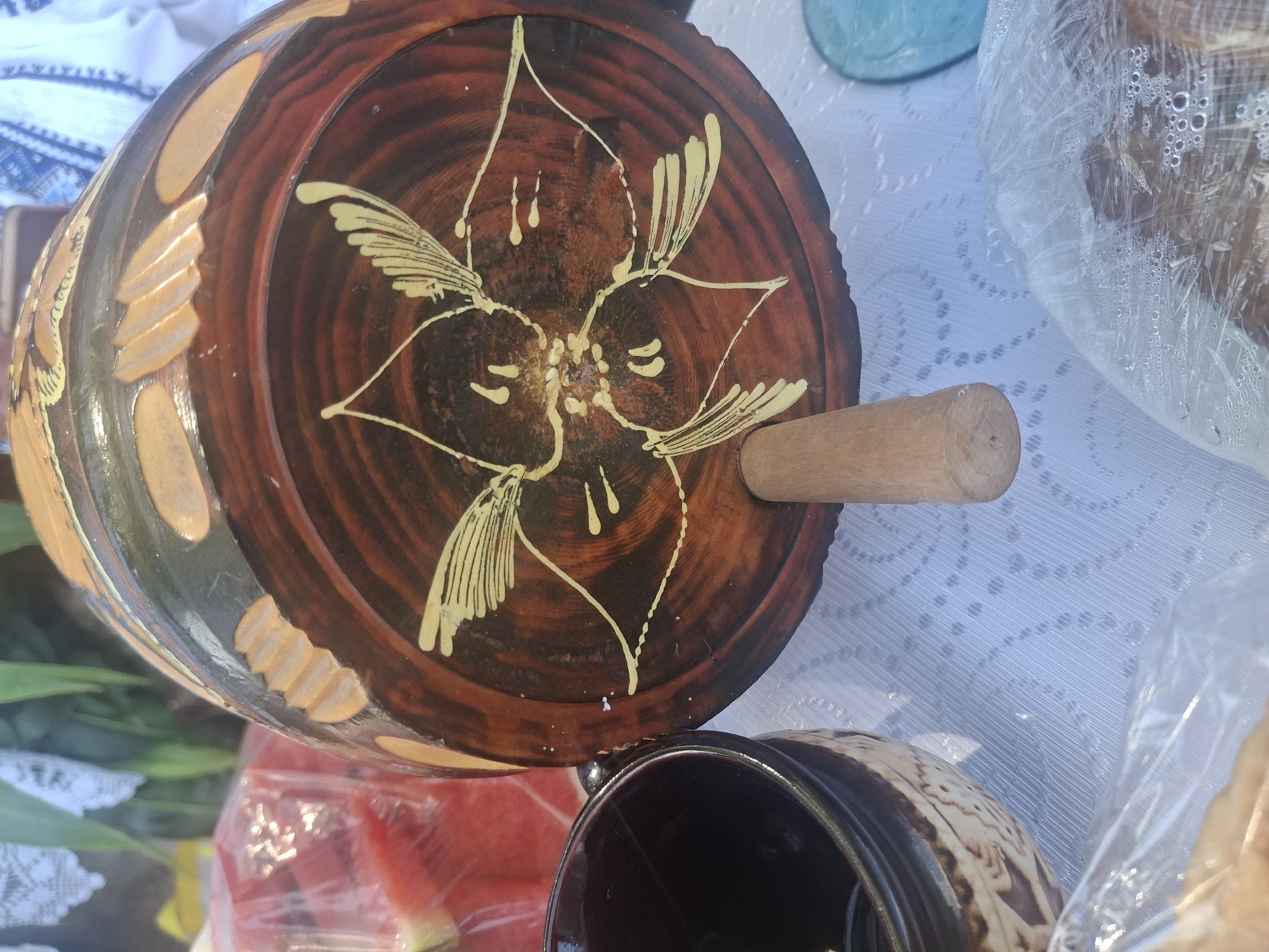 The image depicts a close-up view of traditional Moldovan pottery, likely at an outdoor market or event in Roghi, Moldova. In the foreground, a dark, glazed ceramic mug with an ornate, carved pattern is visible on the left. To its right, a large, wooden, barrel-shaped vessel dominates the scene. The vessel is dark brown with a rich, polished finish and features an intricate, hand-painted yellow design resembling a stylized flower or butterfly on its curved surface. A small, cylindrical wooden peg protrudes from the bottom center of the vessel, serving as a support. The background is softly blurred, revealing hints of other market items: a portion of what appears to be watermelon wrapped in plastic on the far left, and a blue object on the right. The table is covered with a white tablecloth adorned with a delicate lace-like pattern. The overall impression is one of rustic craftsmanship and cultural heritage.