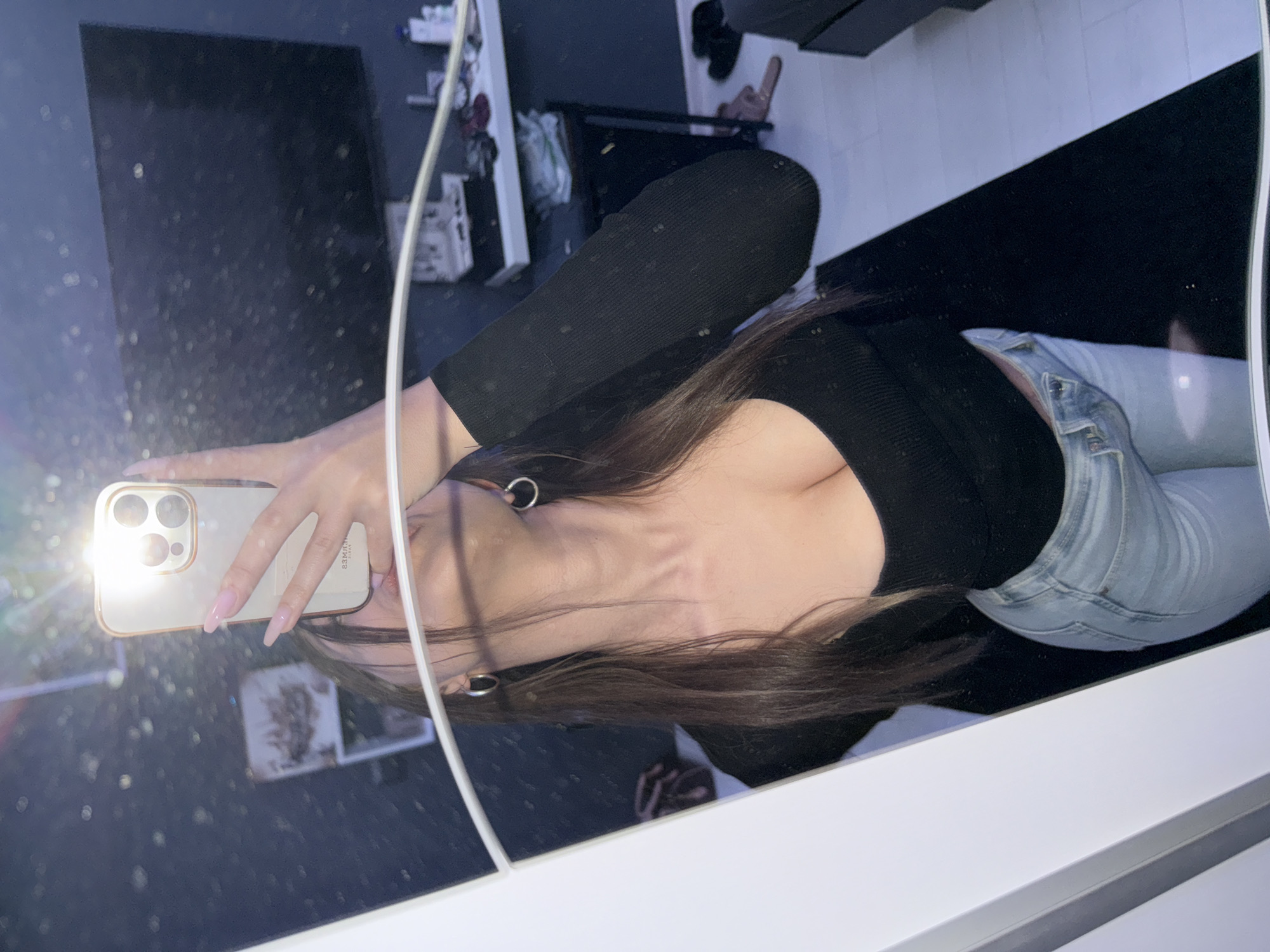 The image shows a woman taking a selfie in a mirror. She is wearing a black long-sleeved top and light blue jeans. She is holding a gold iPhone in her left hand. The flash is on, creating a bright spot on the phone. Her hair is brown and shoulder-length. She is wearing silver hoop earrings. The background is a reflection of a room with a black and white tiled floor. There are various objects reflected in the mirror, including furniture, shoes, and a white cable. The location is not identifiable as Sofia, Bulgaria from the image.