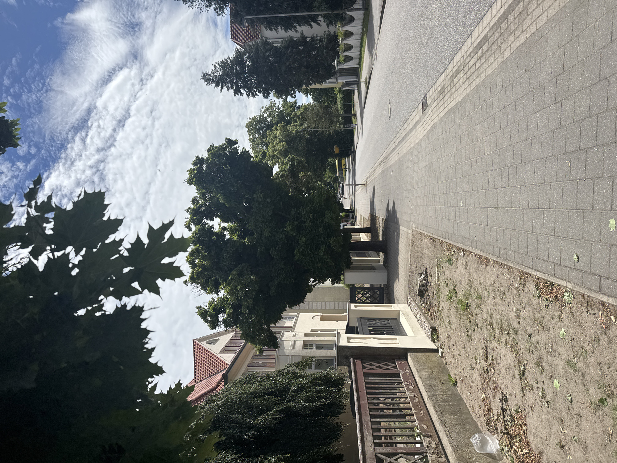The image depicts a street scene in a suburban or residential area, likely in Europe given the architecture. The perspective is from a sidewalk looking down a paved road lined with trees and houses. On the left, there is a weathered wooden fence and a patch of dry, sandy ground with sparse vegetation and a plastic bag. Adjacent to the fence is a cream-colored building with a red tiled roof and dark shutters. Further down the street, the road curves to the right and is flanked by more trees and buildings. A yellow bus is visible in the distance, and a person is walking on the sidewalk. The sky is partly cloudy with white, wispy clouds against a blue backdrop.