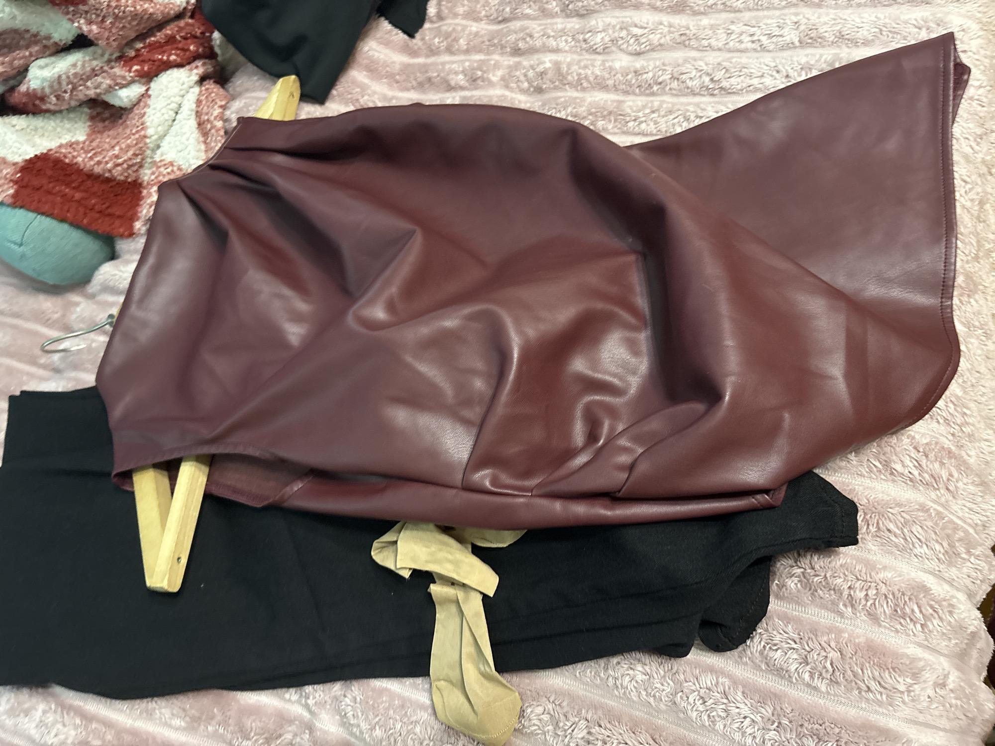 The image shows a close-up view of several items of clothing lying on a textured, pink blanket. A burgundy, possibly faux leather, skirt is draped over a wooden hanger. Underneath the skirt and hanger is a black garment, possibly pants or a skirt, with a light-colored tie detail. To the left, partially visible, is a red and white plaid blanket. The setting appears to be an interior space. There is no visible information indicating the location is Vsevolozhsk, Russia. No people, actions, or interactions are discernible in the image.