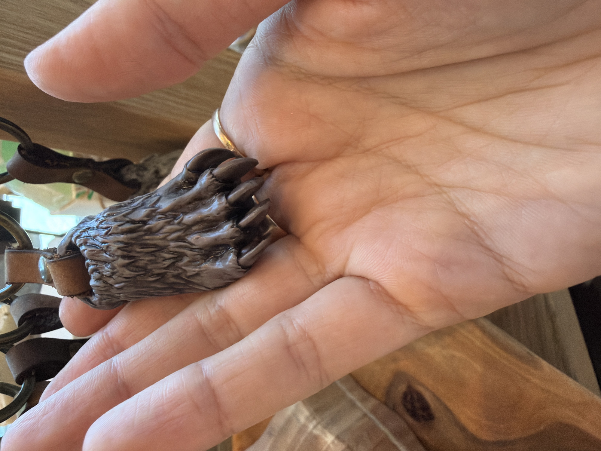 A person holds a dark brown bear claw keychain in their left palm. The hand features a gold ring. The background includes wood surfaces and other parts of the keychain. Location: Cannot be determined from the image.