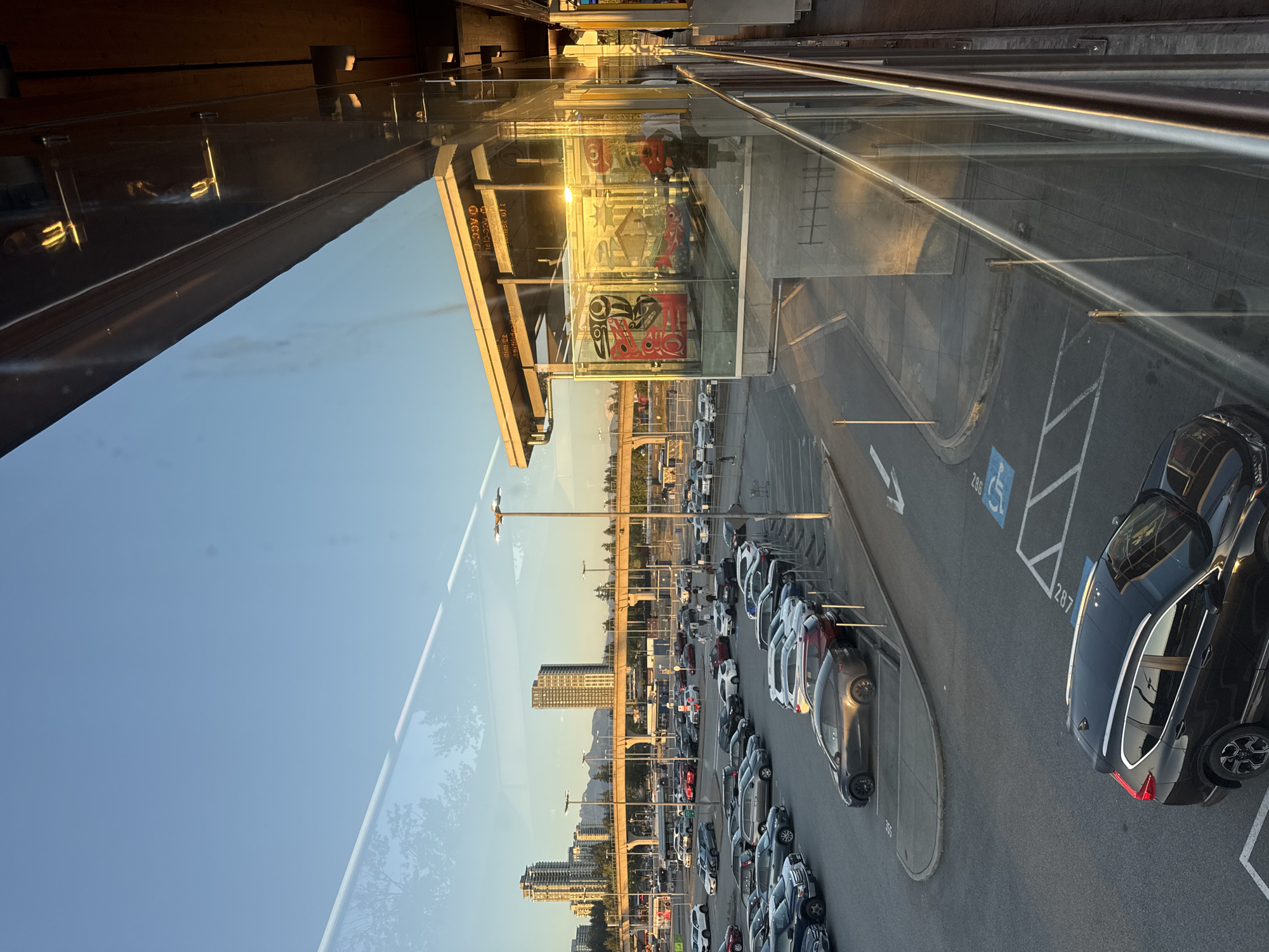 This is an outdoor, eye-level, medium-shot photograph taken from an elevated perspective, likely a covered walkway or a building, looking out over a parking lot towards a city skyline. The scene is illuminated by the warm, low-angled light of either sunrise or sunset.
In the foreground, a modern glass-enclosed structure, possibly a transit station or waiting area, occupies the right side of the frame. The glass reflects the sky, trees, and the interior of the structure, which features what appears to be indigenous artwork displayed on the walls and the faint silhouette of people inside. A metal railing runs along the bottom edge of the frame in front of the glass.
Beyond the glass structure, a large asphalt parking lot is filled with numerous parked cars of various makes and models. Some cars are in designated parking spots, while others are scattered across the lot. The parking lot appears to be quite full.
In the middle ground, a wide elevated roadway or bridge structure with concrete supports spans across the scene, with traffic visible on it. Beyond the roadway, a city skyline with several tall buildings is visible, silhouetted against the clear blue sky. In the far distance, mountains can be seen.
The sky is mostly clear with a few faint contrails. The overall impression is of a modern urban environment in Canada, possibly Coquitlam, Canada, given the architectural style and the presence of mountains, illuminated by the golden hour light. The reflection on the glass creates a layered effect, blending the immediate surroundings with the distant cityscape.