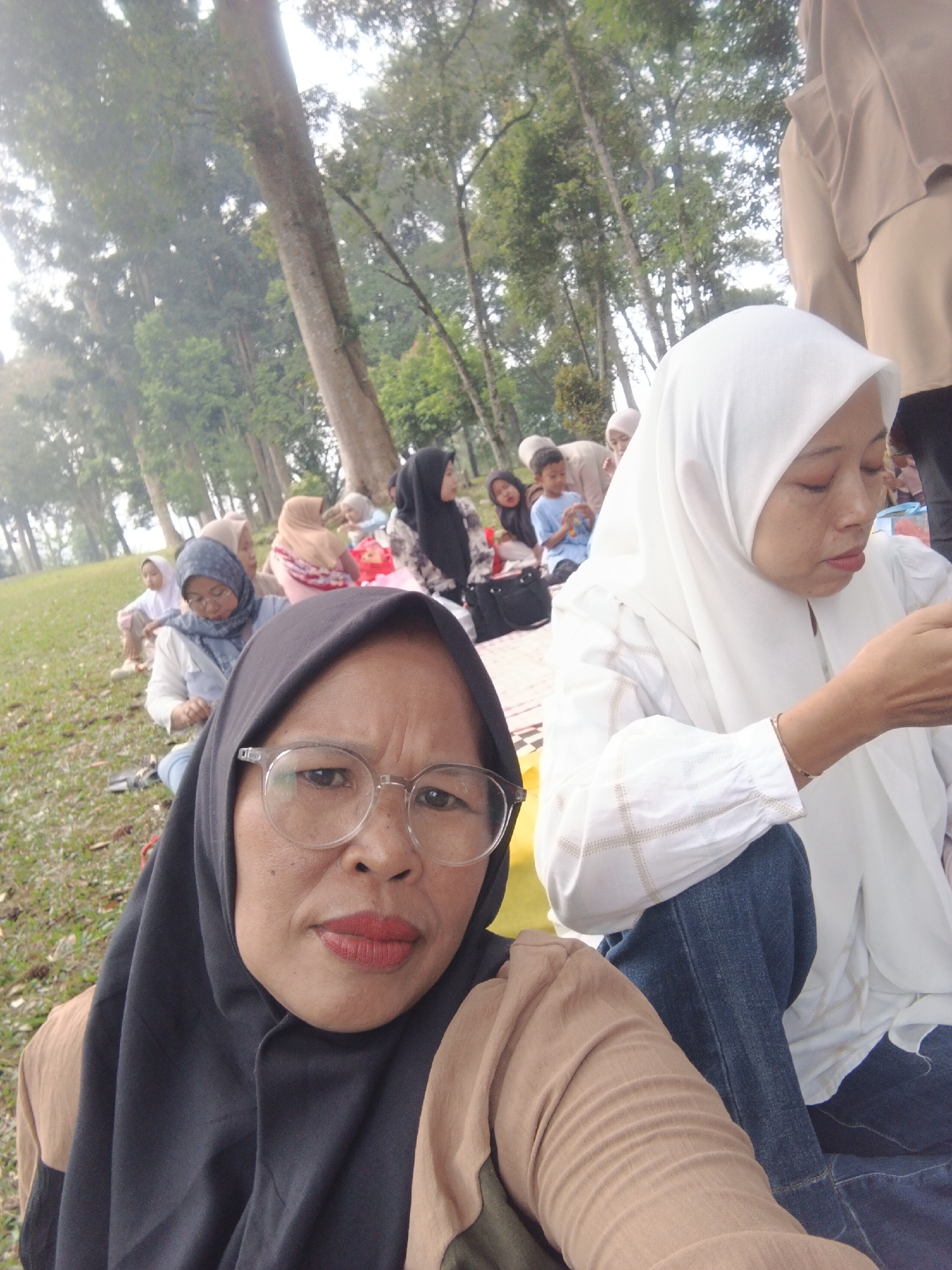 The image shows a group of people picnicking outdoors. In the foreground, a woman wearing a black hijab and glasses is taking a selfie. She is wearing a tan top. To her right, another woman in a white hijab and white shirt is visible, with her eyes closed. Several other people, including children, are seated on the grass in the background, some wearing hijabs. The setting appears to be a park with tall trees and a grassy area. It is unclear if the location is specifically in Sudajaya Girang, Indonesia, as this is not visually confirmed.