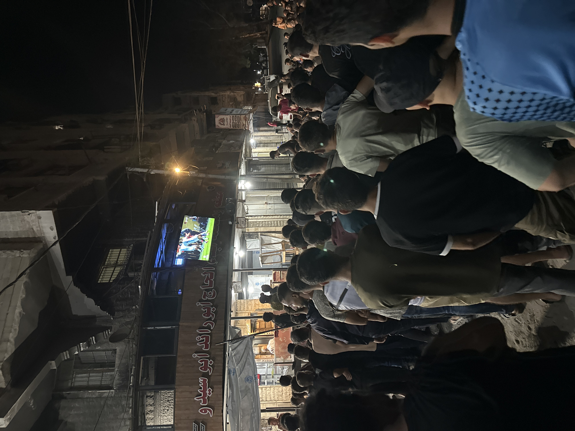 In Gaza City, Palestinian Territories, a crowd of men and boys stand gathered on a dirt street at night, watching a soccer match on a large television screen mounted above a storefront. The screen displays the game with the players in motion, while the spectators are focused on the broadcast. The storefront below the television has Arabic writing on its facade. The surrounding buildings appear aged and weathered, with some showing signs of damage. Streetlights and the glow from the television illuminate the scene, casting long shadows and creating a sense of community as people come together to watch the event. The atmosphere appears to be one of shared interest and engagement.