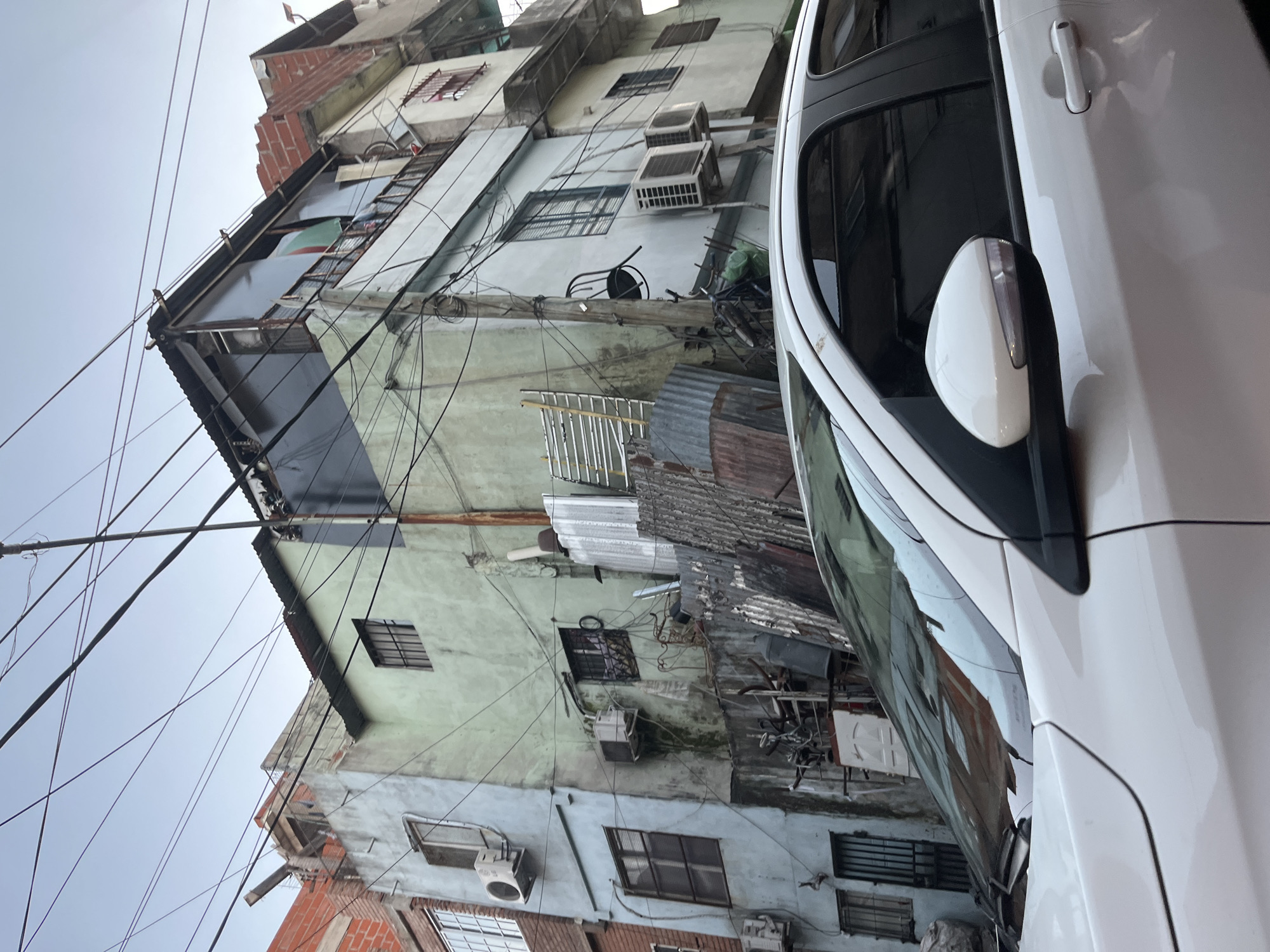 The image captures a dense urban street scene in Buenos Aires, Argentina, looking upwards at a cluster of closely packed multi-story buildings and a parked white car. The buildings, which dominate the left and center of the frame, display a worn appearance with facades in faded light green, light blue, and white, interspersed with sections of exposed red brick. Numerous windows, some secured with bars and others featuring shutters, are visible across the structures. Multiple outdoor air conditioning units are mounted on the building walls.
A complex web of overhead electrical and communication wires crisscrosses the sky, connecting between buildings and poles. Elements of informal construction, such as sections of corrugated metal used for roofs or extensions, are apparent on various levels. A metal ladder leans against one building, and a black-framed chair is perched on a ledge.
On the right side, the front-right portion of a white car is visible, including its side mirror with an integrated turn signal, and the door handle. The car's polished surface reflects the surrounding buildings. The sky above is uniformly light gray and overcast, suggesting a cloudy daytime setting. No people or specific activities are visible, and there is no discernible text. The scene evokes the dense, lived-in character often found in urban informal settlements or older, bustling neighborhoods.
