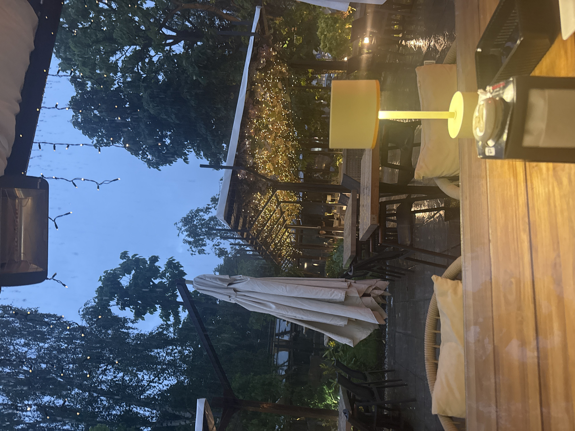 This media file presents an inviting outdoor dining or lounge area, likely a terrace belonging to a cafe or restaurant in Sofia, Bulgaria, captured during dusk or early evening. The scene is bathed in a warm, cozy light from numerous illuminated fixtures.
The foreground, on the right side of the image (when correctly oriented), features a wooden tabletop hosting a modern table lamp with a soft yellow shade. Next to it, a small electronic device or remote control is visible, along with a faint tag displaying "www.artemide.it", indicating the brand of the lamps. A table number, possibly "19", is also discernible. Part of a wicker chair with a light-colored cushion is visible at the edge of the frame.
The midground is dominated by additional wicker chairs with cushions and a large, neatly folded beige patio umbrella. A prominent pergola or canopy structure is extensively draped with countless warm string lights, creating a magical and festive atmosphere.
In the background, dark-foliaged trees stand out against a light blue, twilight sky. More string lights are strung among the trees or overhead. The presence of small reflective specks on darker surfaces, especially visible on the top left, suggests recent precipitation or a damp environment. To the far left, a patio heater or outdoor lighting fixture is partially visible.
No people are present, suggesting a tranquil moment, perhaps before opening or after closing hours. The soft, ambient lighting and comfortable seating evoke a serene and relaxed mood, ideal for an evening gathering.