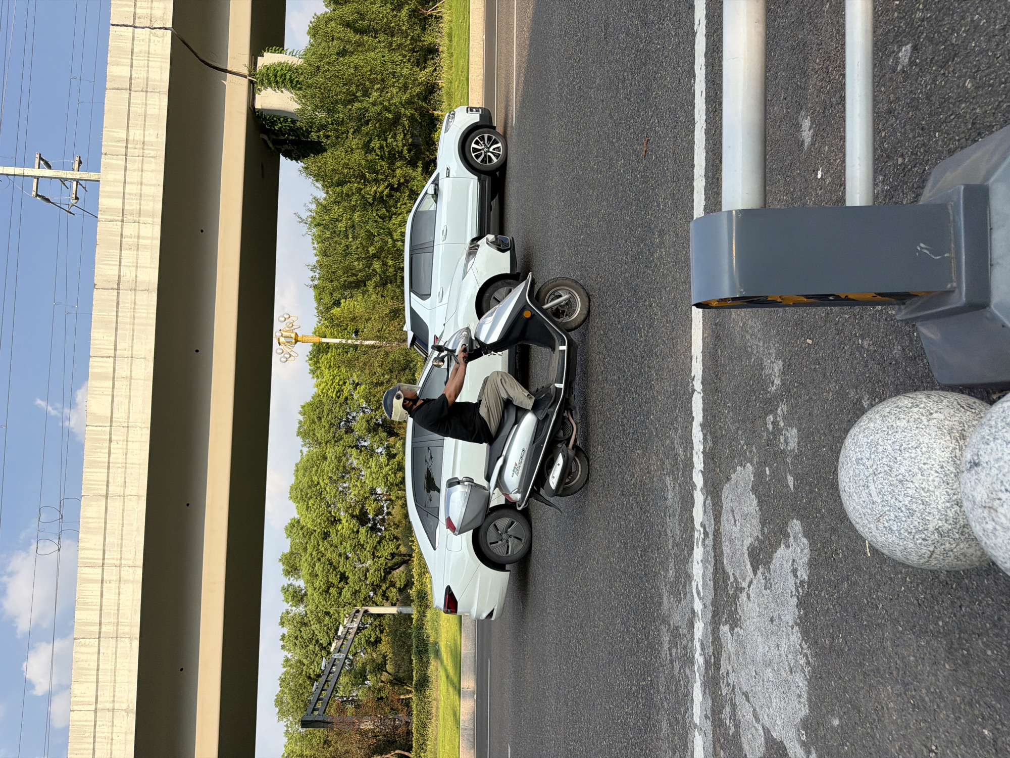 A man wearing a black shirt, khaki pants, and a helmet with a clear visor rides a silver scooter on a road under a concrete overpass. The scooter has a silver storage box on the back. A white sedan is to the left of the scooter, and a white SUV is to the right. Trees line the side of the road behind the vehicles, and power lines are visible above the overpass. The scene appears to be in a city in Jiangjin, China. The road is asphalt with white lane markings. In the foreground, part of a gray traffic barrier and two spherical, granite bollards are visible.