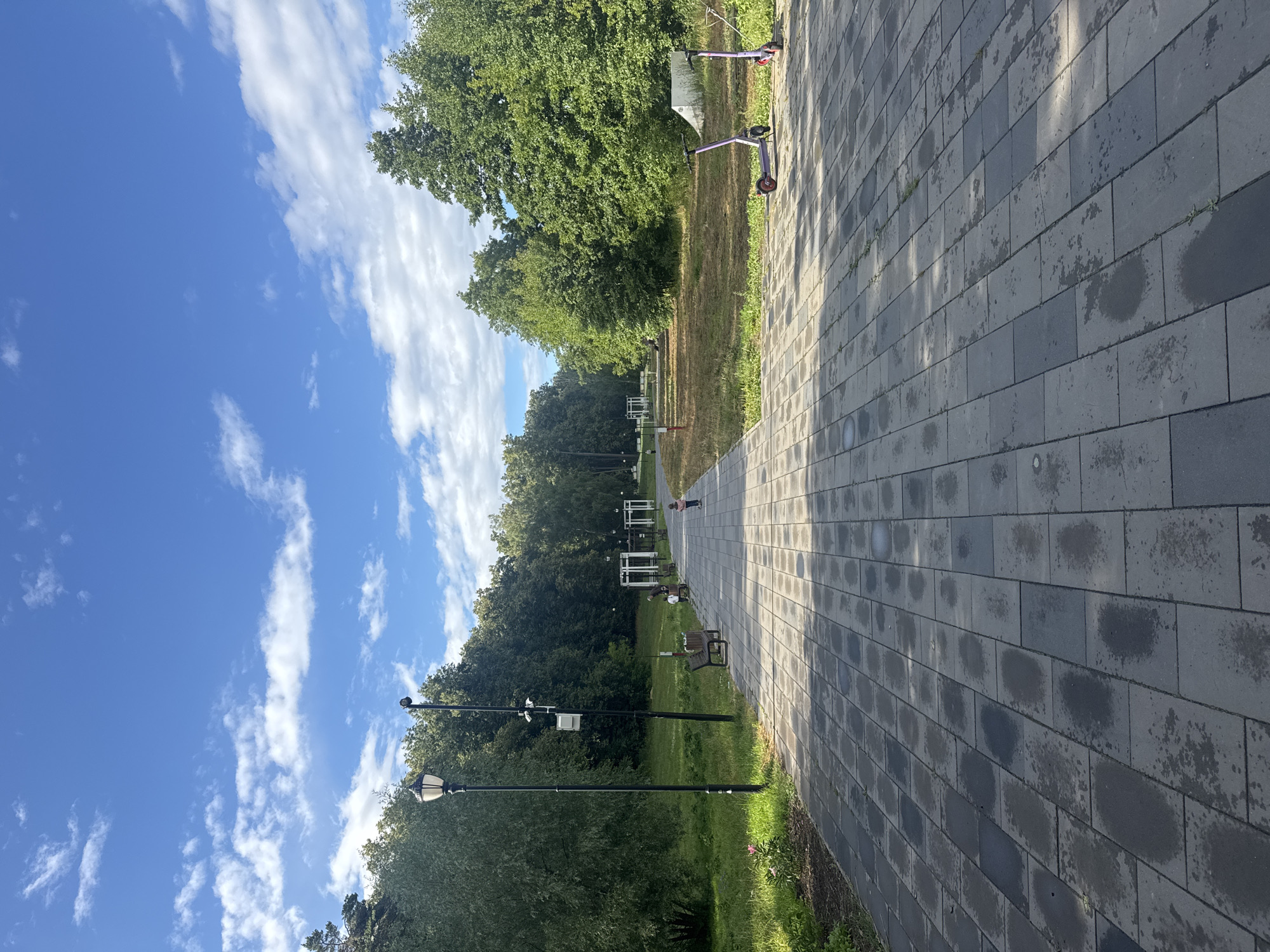 The image depicts a paved park walkway in Sarov, Russia, under a bright blue sky with scattered white clouds. The walkway is made of rectangular grey pavers, some of which appear wet, suggesting recent rain or watering. To the left of the walkway is a grassy area with trees, a lamppost, and a bench. Further down the walkway, there is a child on a scooter and another child standing near a bench with an adult. To the right of the walkway, more trees and a grassy area are visible, with two purple electric scooters parked. In the background, there are white structures that resemble gazebos or pergolas, and a dense line of trees forming the park's boundary. The overall scene is one of a well-maintained urban park on a pleasant day.