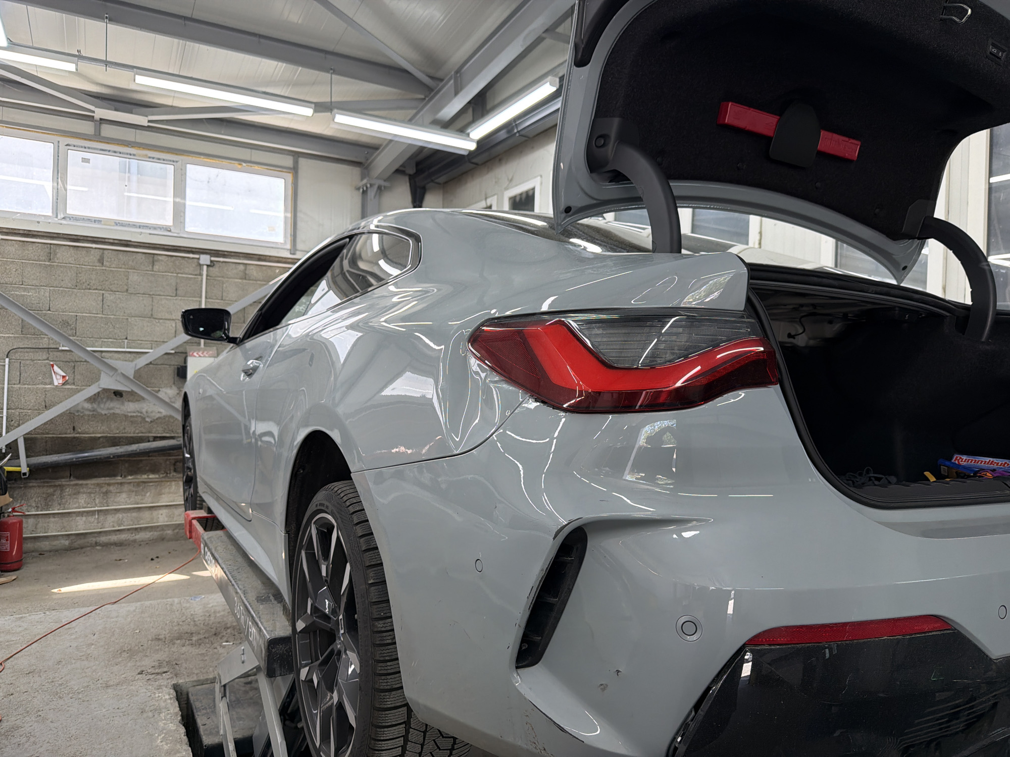 A gray BMW coupe is raised on a lift inside a repair shop. The car's trunk is open, revealing a dark interior. The exterior of the car appears to have been damaged on the right rear quarter panel and on the right rear bumper with multiple scratches. The shop has a brick wall in the background with three windows that offer outside visibility, revealing it is daytime. There is additional lighting from above that illuminates the area. A red fire extinguisher can be seen next to some steps. The location is Burgas, Bulgaria.