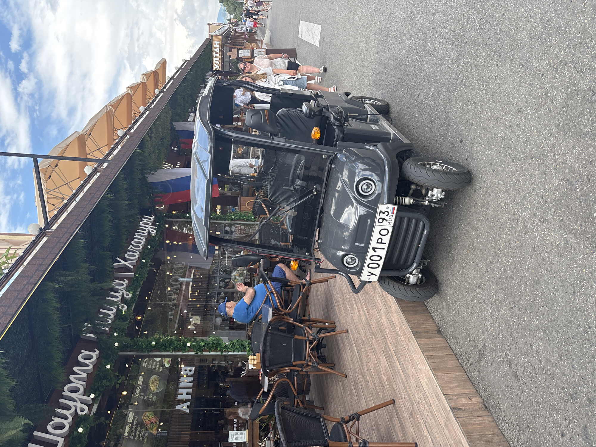 The image depicts a street scene in Sochi, Russia, near a cafe with outdoor seating. In the foreground, a small, dark grey electric vehicle with a license plate reading "Y001PO93" is parked on the asphalt. The vehicle has a canopy and open sides. To the left of the vehicle, there is a wooden deck with several empty outdoor tables and chairs. A man in a blue t-shirt and shorts is seated at one of these tables, looking towards his left. The cafe building behind the tables has a large glass facade decorated with artificial greenery and string lights. Signs with Russian text are visible on the cafe's exterior. In the background, several people are walking along the street, and other buildings and trees can be seen. The sky is cloudy.