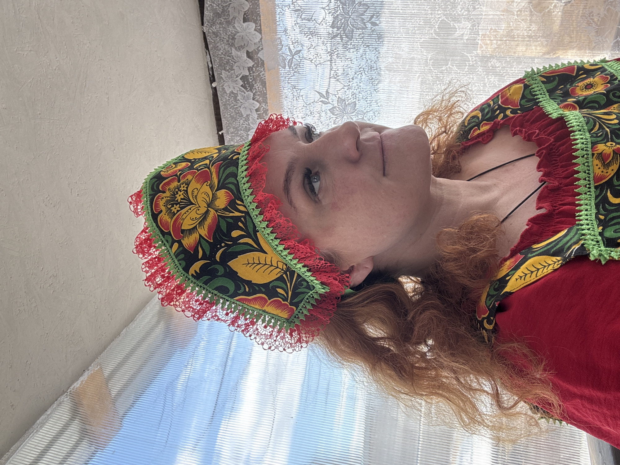 The image shows a woman with light skin and red, curly hair wearing a traditional Russian dress and head covering with floral patterns and red lace. The dress is primarily red with similar floral details around the neckline. The background consists of a window with white floral lace curtains, giving the impression of natural light. The setting appears to be indoors, possibly a home or studio. There is no visible context to confirm the location as Chaikovsky, Russia.