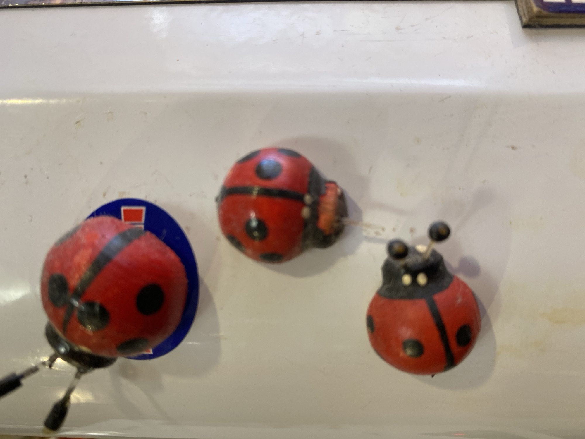 The image shows three decorative ladybugs attached to a white surface, likely a refrigerator or a similar appliance. The ladybugs are red with black spots and have protruding antennae. The top ladybug has a blue circular backing with a red square in the center. The white surface appears slightly worn and has some smudges or dirt marks. The lighting suggests an indoor environment. The image does not contain information about the city of Novaya Usman', Russia.