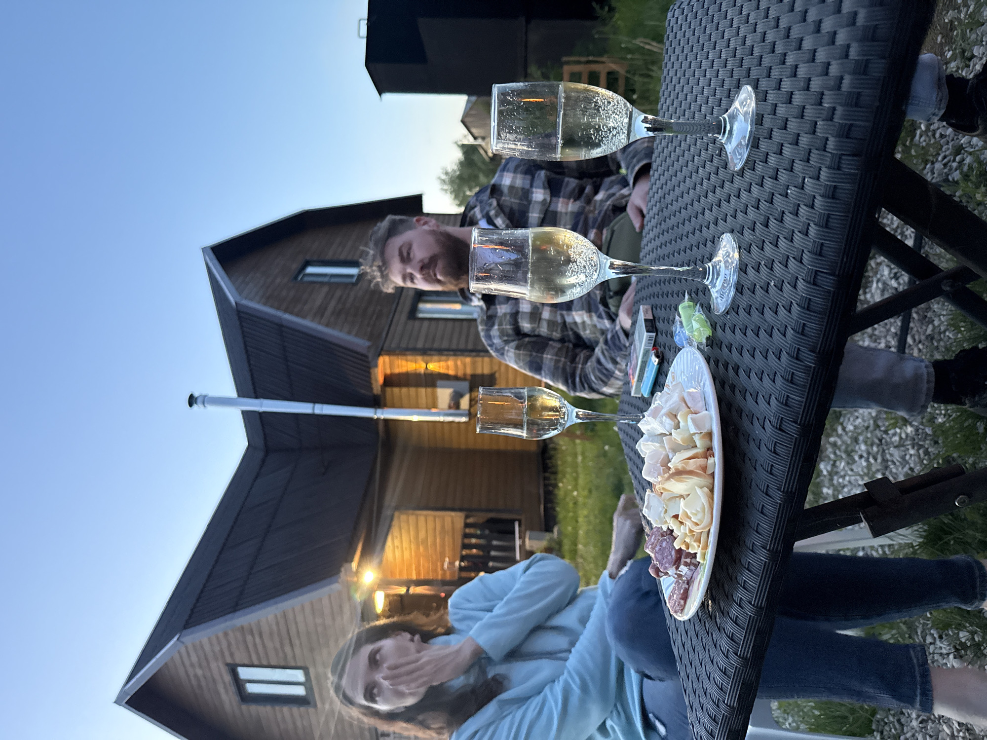 A medium shot captures two individuals seated at a black wicker table outdoors. A man with a beard, wearing a plaid shirt, is looking at the camera. A woman with long brown hair, wearing a light blue sweatshirt and jeans, is sitting to his left, and her hand is in front of her mouth. Three glasses of sparkling wine are placed on the table, along with a plate of cheese and charcuterie, lighters, and a cigarette pack. In the background, a wooden house with dark roofing is visible, illuminated with warm light. The setting appears to be a yard or garden, with green grass visible between the table and the house. The location is Khutyn', Russia.