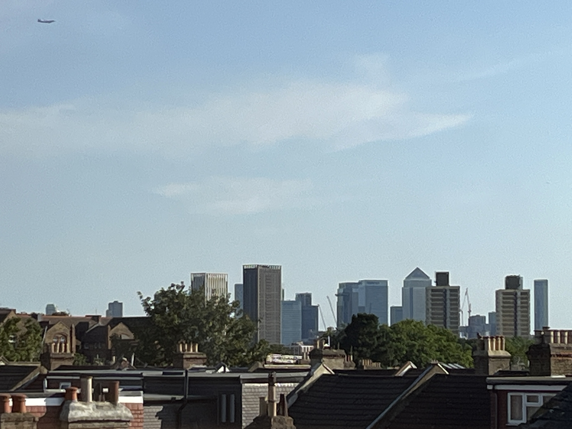 The image shows a distant view of the Canary Wharf skyline in London, United Kingdom, seen over a row of residential rooftops. In the upper left corner of the sky, a small, dark silhouette of an airplane is visible. The foreground is dominated by the pitched roofs of terraced houses, with chimneys of various sizes and designs protruding from them. The middle ground features trees, some with green foliage, interspersed with the architecture of residential buildings. The background is filled with the modern skyscrapers of Canary Wharf, characterized by their varied heights and architectural styles. The sky is clear and light blue, with some faint white clouds. The overall impression is a contrast between traditional urban housing and a modern business district.