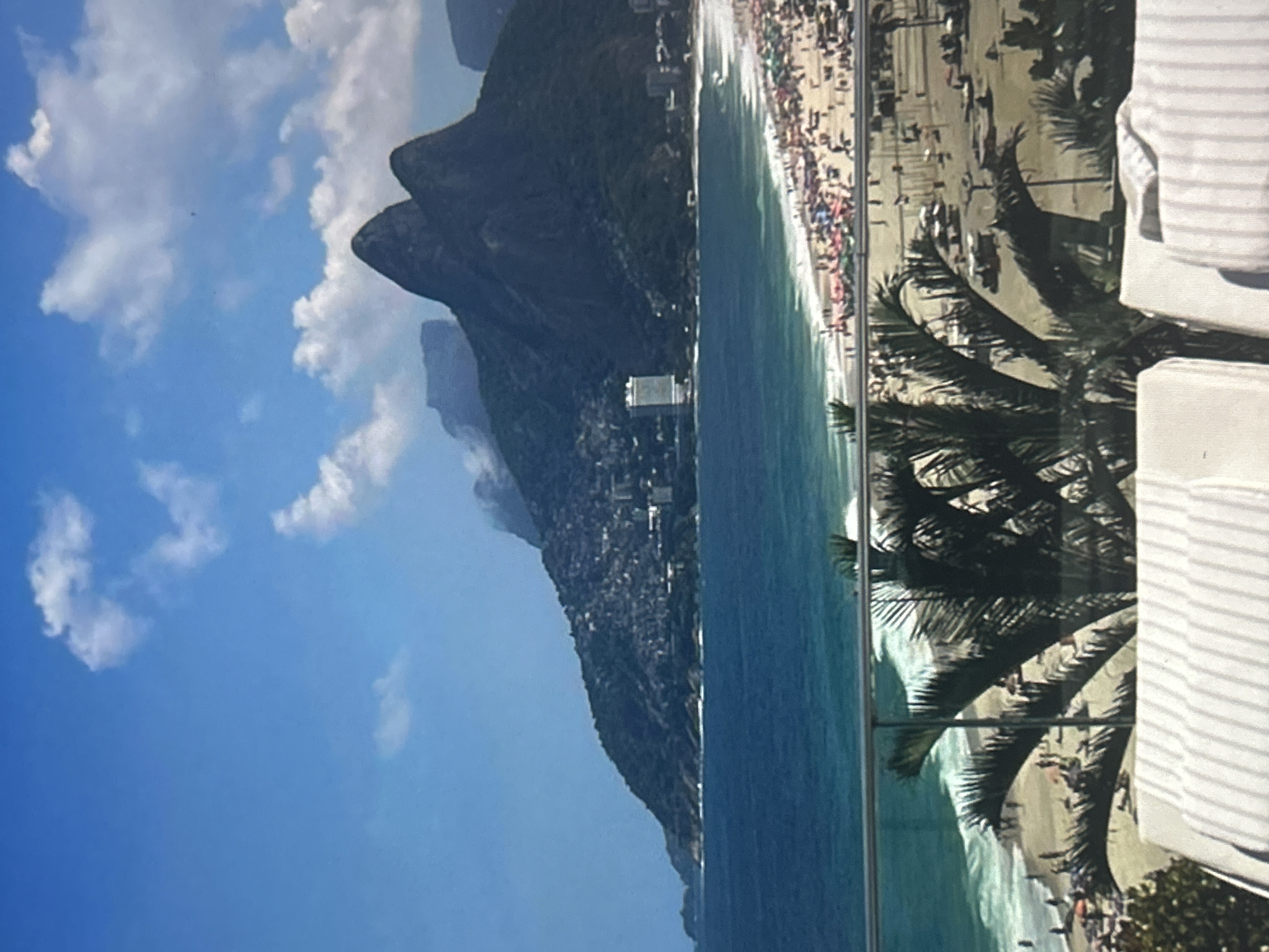 This is a landscape photograph taken from an elevated perspective, looking out over a bustling beach and a large, mountainous landmass. The scene is dominated by a bright blue sky with scattered white clouds. The ocean is a vibrant turquoise, with white waves crashing onto a sandy beach. The beach is crowded with people, suggesting a sunny and warm day, likely midday given the brightness of the light.
The mountainous terrain is covered in lush green vegetation and dotted with buildings, indicating a populated coastal area. The prominent peak of the mountain resembles a human profile. Palm trees with distinct fronds are visible in the foreground, partially obscuring the view of the beach and suggesting a tropical or subtropical climate. A glass barrier is present in the foreground, possibly part of a balcony or viewing platform, with a striped fabric draped over a portion of it on the right side of the frame.
The overall mood is one of leisure and vibrant activity, with the clear sky and crowded beach conveying a sense of a popular tourist destination. There is no visible text in the image.