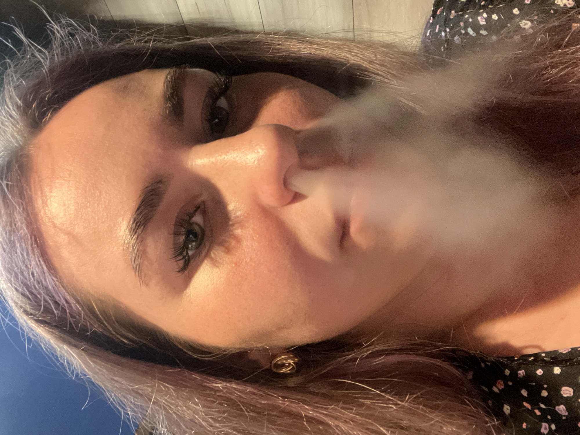 A close-up, angled portrait shows a woman exhaling a thick cloud of white smoke. Her face is fair, with pronounced eyelashes and slightly arched eyebrows. She has a gold earring visible on her left ear. Her hair is a mix of dark brown and light purple-gray. She's wearing a dark-colored top with a pattern of small white and pink shapes. The background is blurred, suggesting a shallow depth of field. There's a hint of blue at the bottom left and a light-colored wall at the top. There is no indication of this image being related to the city of Chaikovsky, Russia.