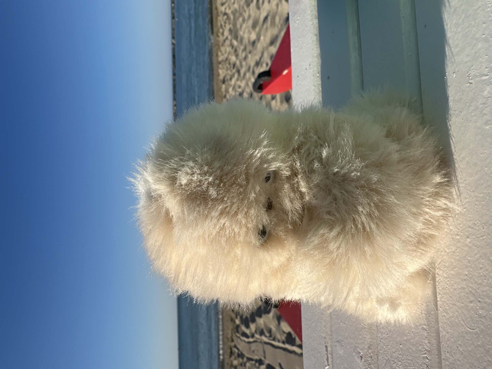 A fluffy, cream-colored stuffed animal is positioned on a white wooden surface in the foreground, obscuring most of the background. The stuffed animal has two small, dark eyes visible through its fur. Behind it, a sandy beach stretches towards the sea. In the distance, the blue water meets the horizon under a clear blue sky. Two red objects, possibly beach chairs or rescue equipment, are partially visible on the sand. The scene appears to be a sunny day at the beach, likely in Pesaro, Italy, given the context of the prompt.