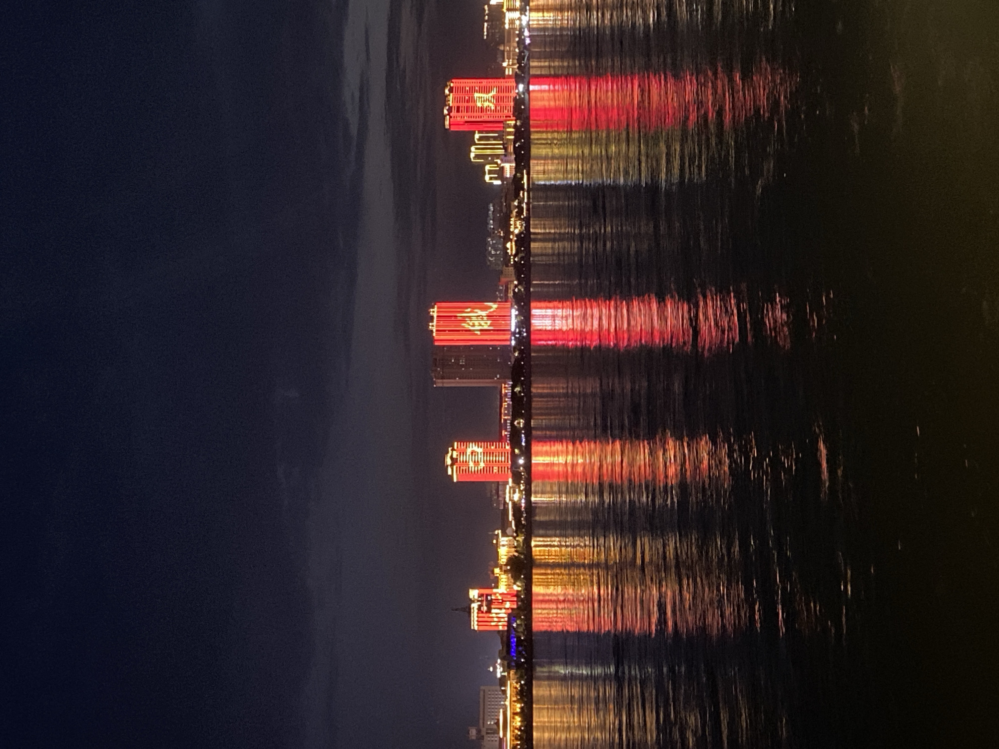 The image displays a night scene of the city of Blagoveshchensk, Russia, as viewed from across the Amur River. The foreground is dominated by the dark, rippling water of the river, reflecting the bright lights of the cityscape. The background features a line of illuminated buildings, some of which are tall, modern structures adorned with red and yellow lights. The buildings appear to be illuminated with large Chinese characters, suggesting a significant presence of Chinese culture or business in the city. The sky above is a deep, dark blue, with hints of lighter clouds towards the horizon. The overall impression is one of a vibrant, modern city at night, with a strong visual connection to China across the river.
