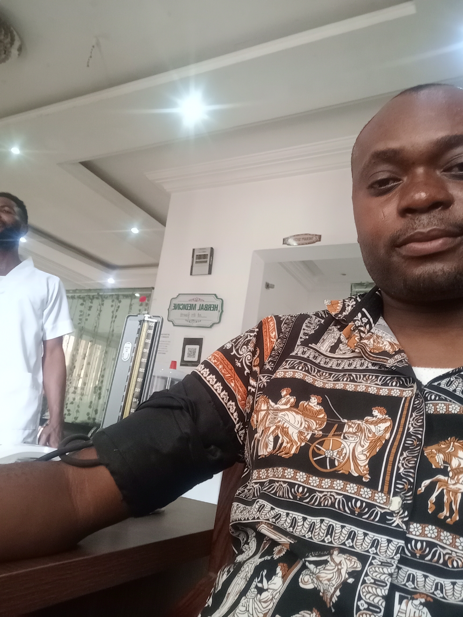 The image appears to be a selfie taken indoors, possibly in a medical or health-related setting. A man is in the foreground, wearing a patterned shirt, with his upper arm and a blood pressure cuff visible on a table. Another man is in the background wearing a white shirt. A medical device, likely a sphygmomanometer, is visible near the man in the background. A sign that reads "HERBAL MEDICINE" (mirrored) is visible in the background. The location appears to be in Gwarinpa, Nigeria.