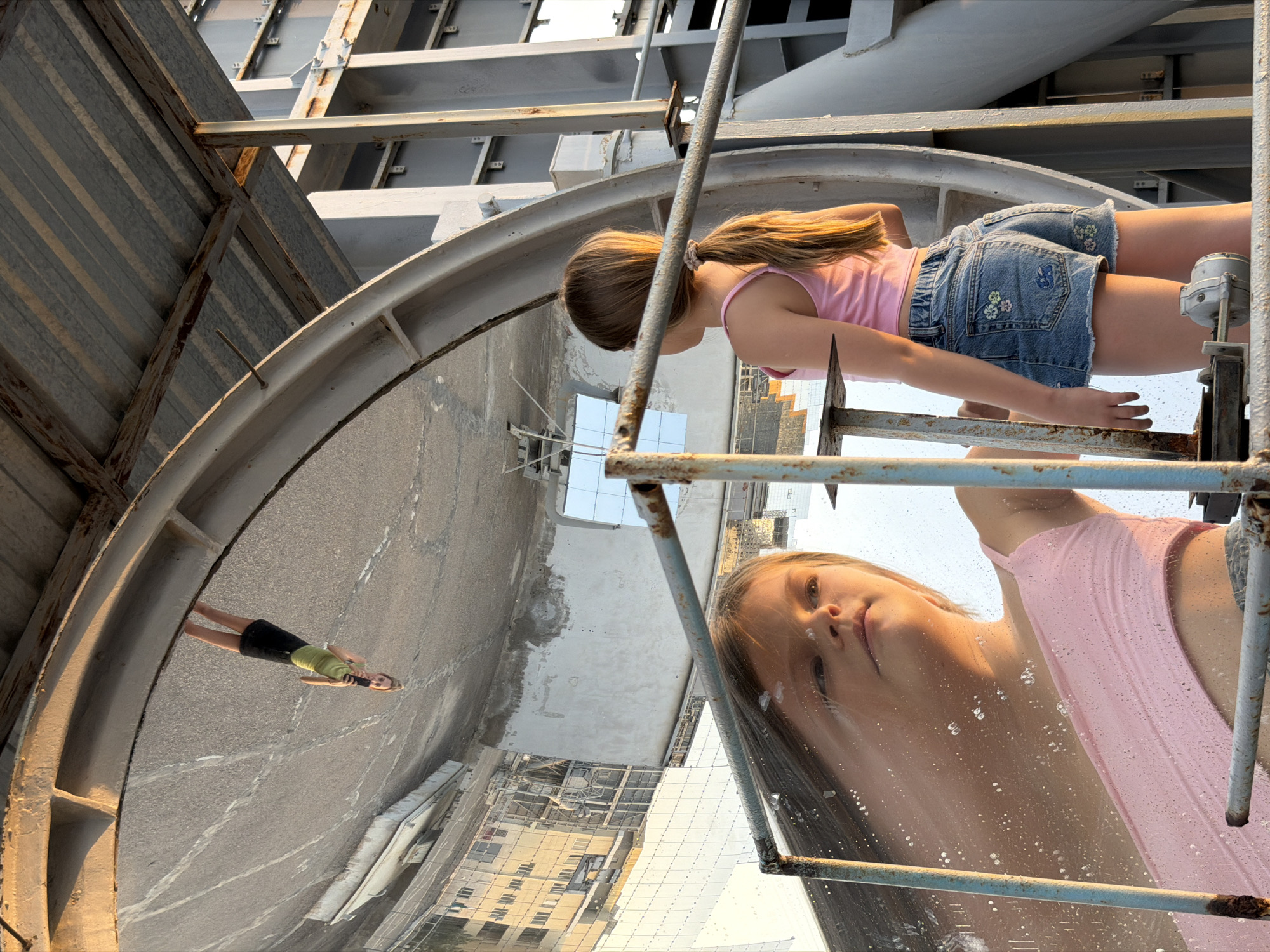 In the city of Hisarak, Uzbekistan, a young girl with blonde hair pulled back in a ponytail stands facing away from the camera, wearing a pink tank top and denim shorts adorned with floral embroidery. She is positioned in front of a large, curved, reflective surface that mirrors the sky and surrounding structures, including a distant building. Her reflection in the surface shows her upside down, wearing the same outfit, and holding a mobile phone. The scene takes place outdoors, with industrial-like metal structures framing the reflective surface and the girl. The lighting suggests it is late afternoon or early evening, with warm sunlight casting long shadows and highlighting the textures of the metal and the girl's hair. The reflective surface appears to be part of a solar concentrator, given its shape and reflective properties, common in areas like Hisarak which have historically utilized such technology for research.