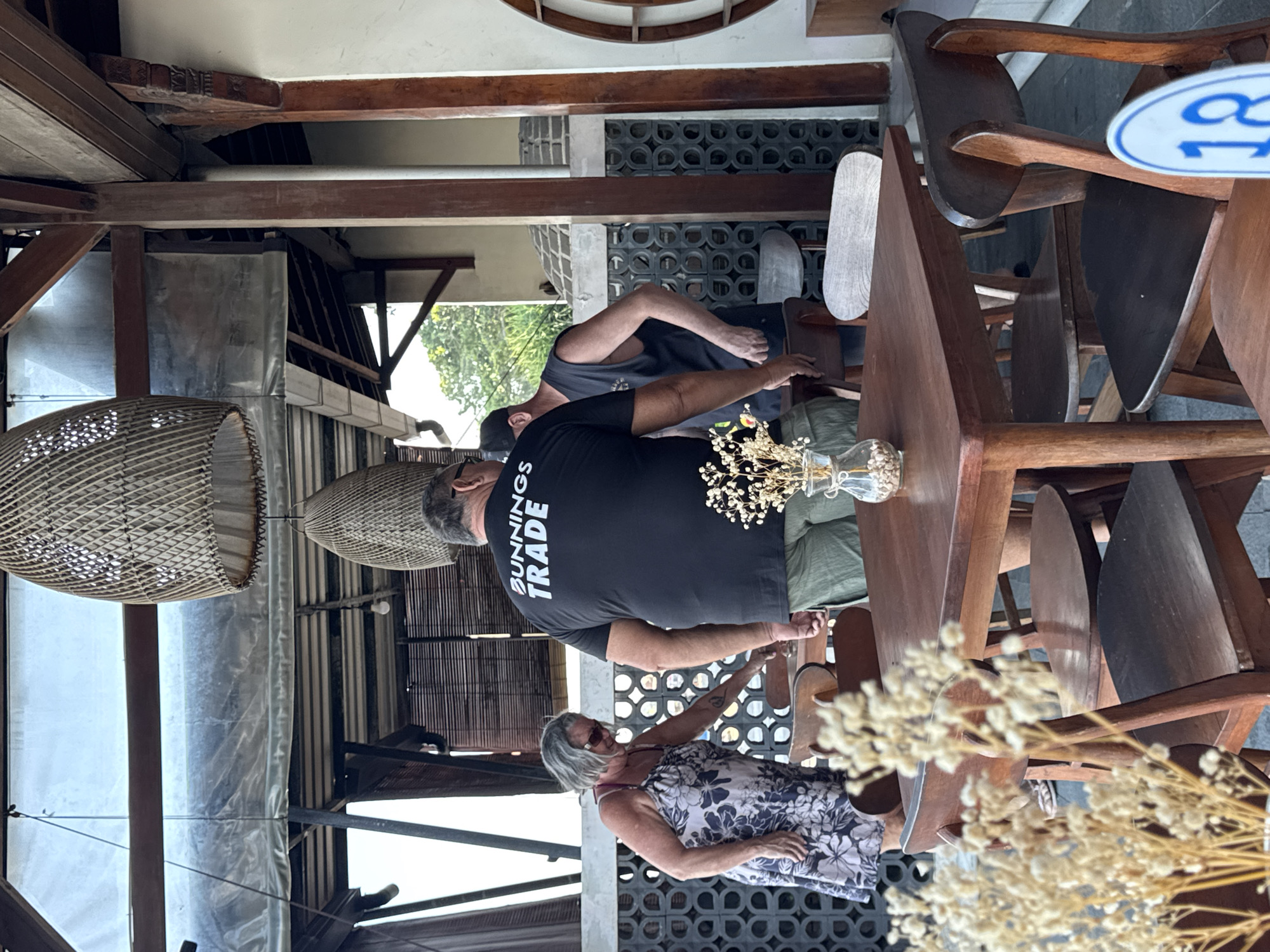 The image captures a casual, semi-outdoor dining or gathering space, likely in a restaurant or cafe in Kuta, Indonesia. The scene is bright, suggesting daytime.
In the foreground and midground, three individuals are visible. A man with grey hair and sunglasses, wearing a black t-shirt with "BUNNINGS TRADE" printed in white on the back, stands facing away from the camera. He appears to be interacting with another man, partially visible, who is wearing a dark tank top. To the right, a woman with grey hair and sunglasses, dressed in a white sundress with a dark floral pattern, stands near a wooden table, looking towards the left.
The setting features rustic wooden furniture, including several tables and chairs, some stacked. On one table in the foreground, a clear glass vase holds dried, light-colored flowers. Overhead, two large, intricately woven light fixtures hang from a wooden beam ceiling. The background reveals architectural details such as decorative concrete blocks forming a wall, more wooden beams, and what appears to be a clear plastic or vinyl sheet acting as a screen. Green foliage is visible outside, indicating an open-air environment. A round blue and white sign, partially visible, displays the letters "CO" and the number "18". The overall atmosphere seems relaxed and informal.