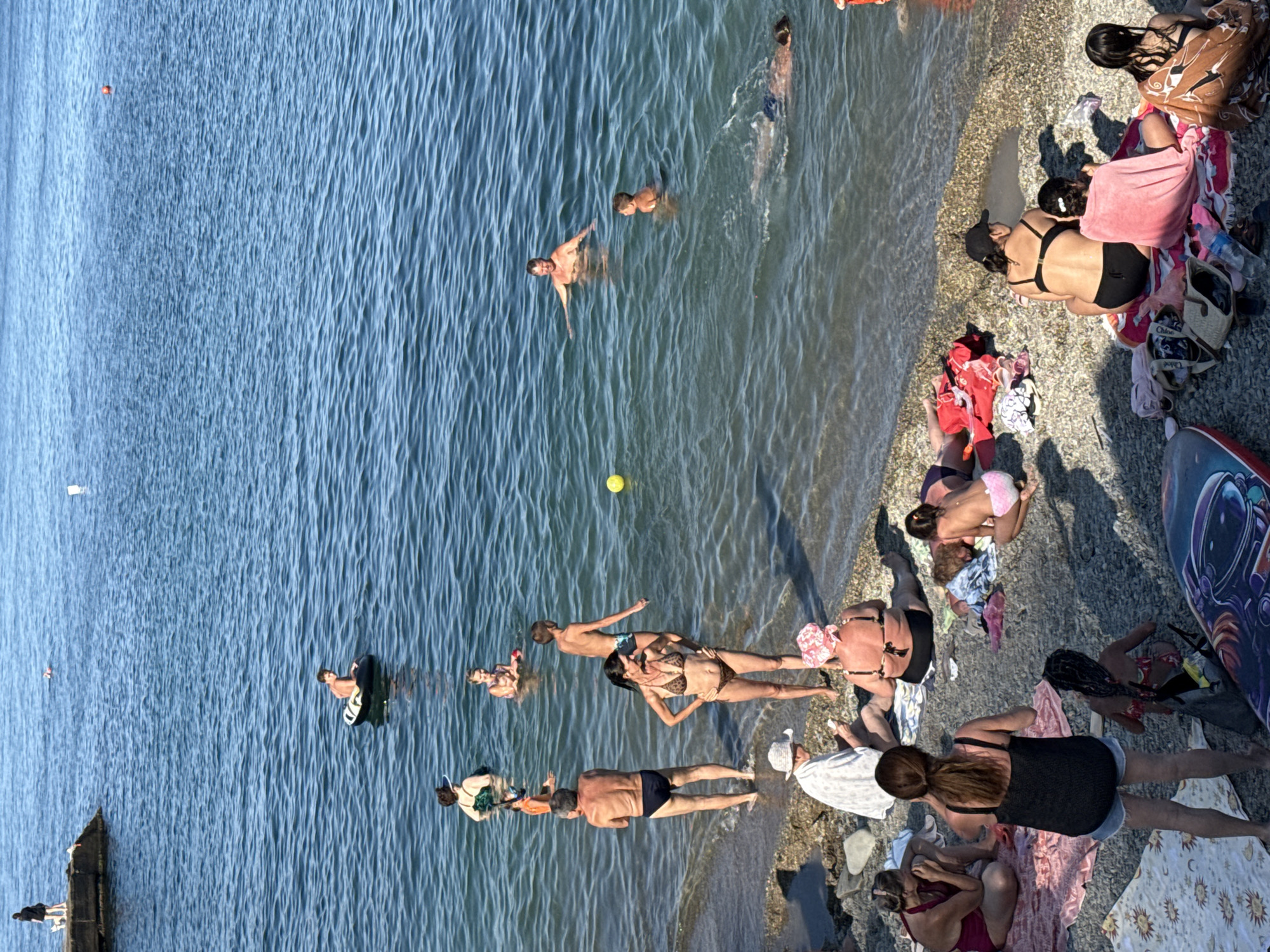 The image depicts a sunny day at a pebble beach in Partenit, Ukraine. Several people are engaged in various activities related to a beach outing. In the water, a man is swimming, while a boy is partially submerged, and another person is visible further out. A yellow ball is floating near the shore. On the beach, people are sitting and lying on towels, some are wearing swimwear, and one person is holding a phone. The background shows the blue sea under a clear sky.