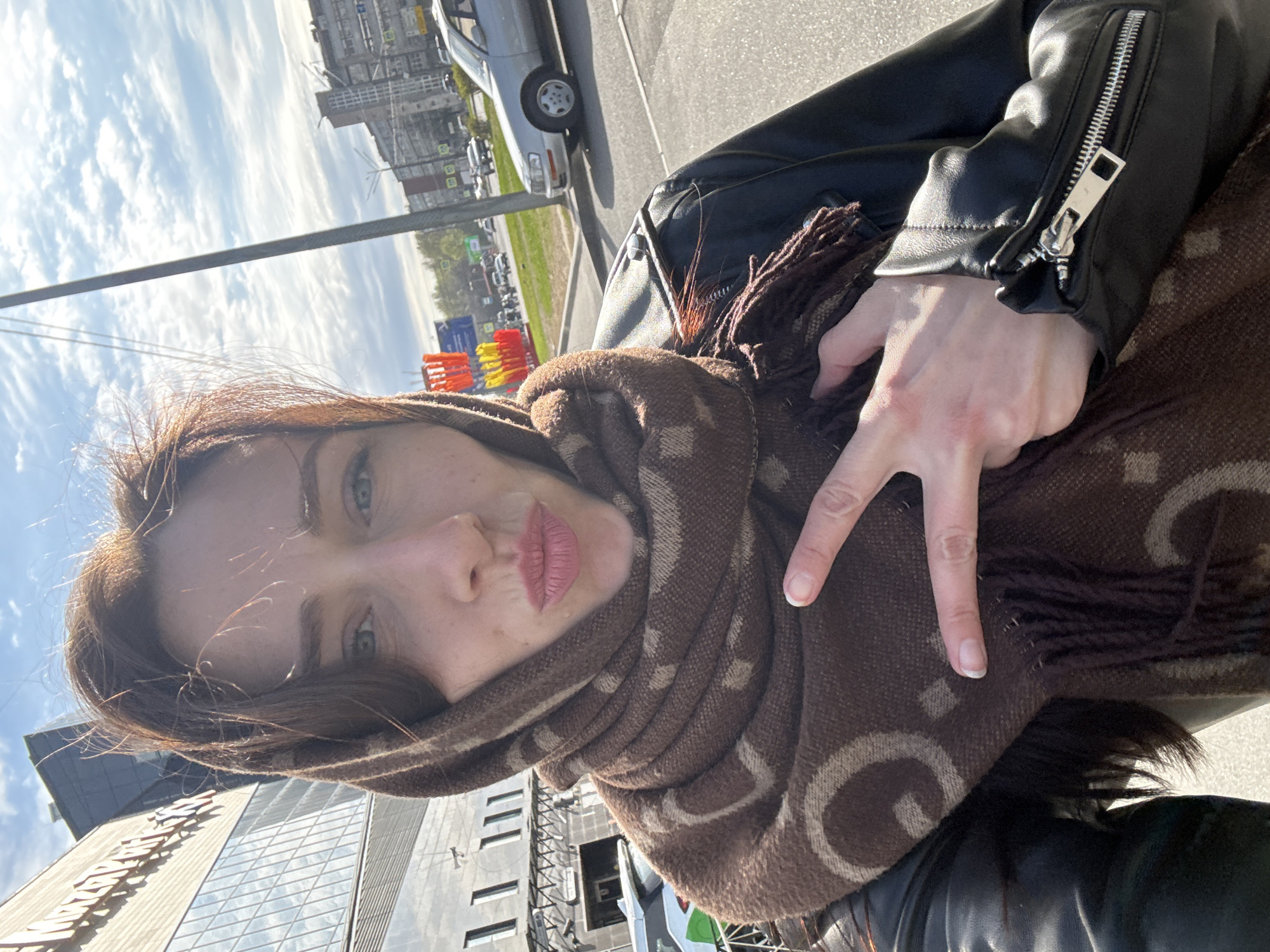 A woman takes a selfie in what appears to be Saint Petersburg, Russia. She wears a black leather jacket and a brown patterned scarf wrapped around her head and neck. Her hand is raised, forming a "peace" sign. The cityscape includes modern buildings with glass facades and a car parked on the street. The sky is partly cloudy, suggesting daylight hours. In the background, traffic barriers, painted in bright orange, red, yellow, and blue can be seen.