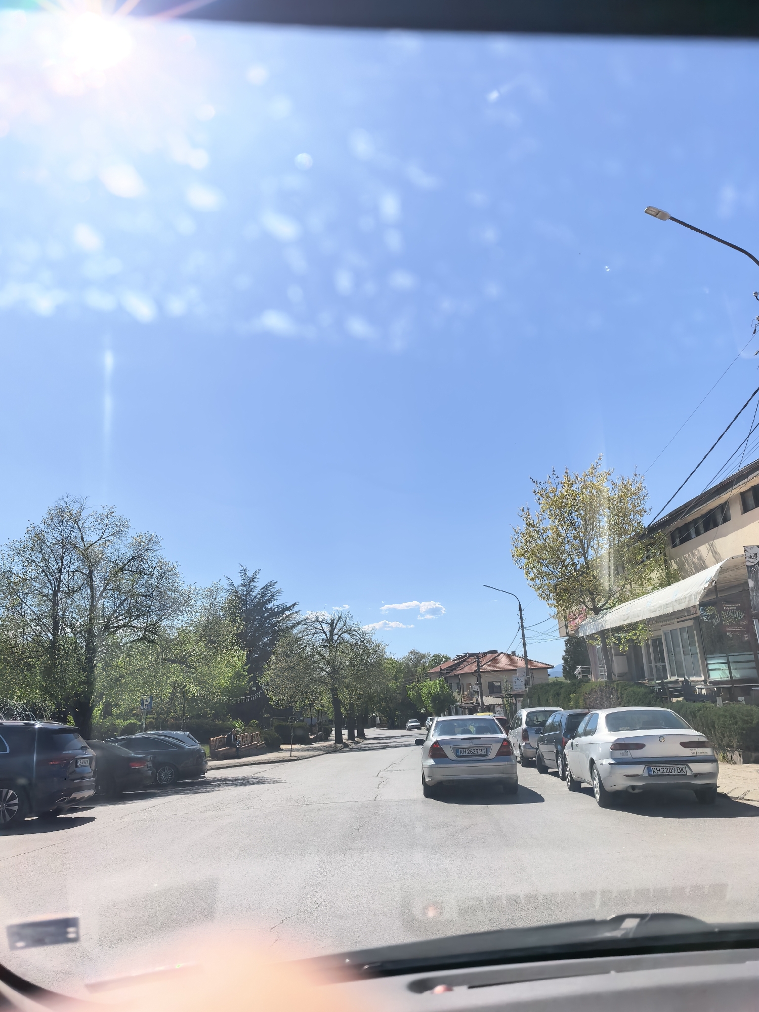 The image shows a street view in Sapareva Banya, Bulgaria, taken from inside a vehicle. The day is sunny with a bright blue sky and a few scattered clouds. The road is paved with asphalt and has some cracks. Several cars are parked on both sides of the street. In the foreground, the dashboard of the vehicle is visible. In the middle ground, a silver Mercedes-Benz with license plate KH 2629 BT, a silver van, a gray vehicle, and a silver Alfa Romeo with license plate KH 2289 BK are visible, all parked in line along the street. On the left side of the street, trees line the road. On the right side, there is a building with an awning and what appears to be a store or restaurant. A streetlight is positioned along the right side of the street. Other houses and buildings are visible in the background. No people are clearly visible in the image.