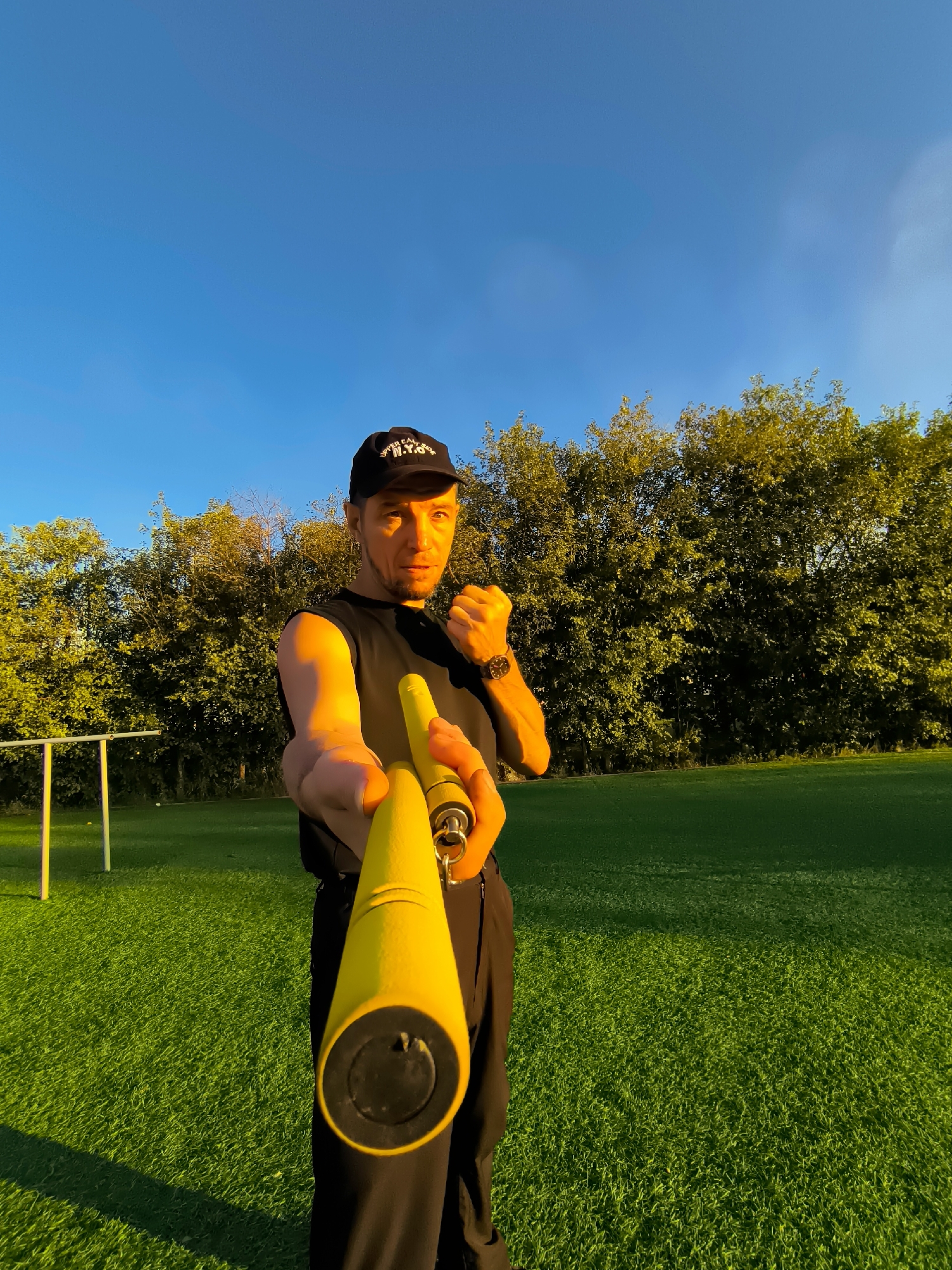 A man is standing on a bright green grassy field with a clear blue sky overhead. He is wearing a black sleeveless shirt, a black baseball cap, and black pants. He is holding a yellow training tool in his right hand, extending it towards the camera. The man is looking directly at the camera with a serious expression and his left fist is clenched. Behind him, there is a line of trees with green foliage, and in the distance, a white sports goal structure is visible on the left side of the frame. The lighting suggests it is late afternoon or early evening, with warm golden light illuminating the scene.