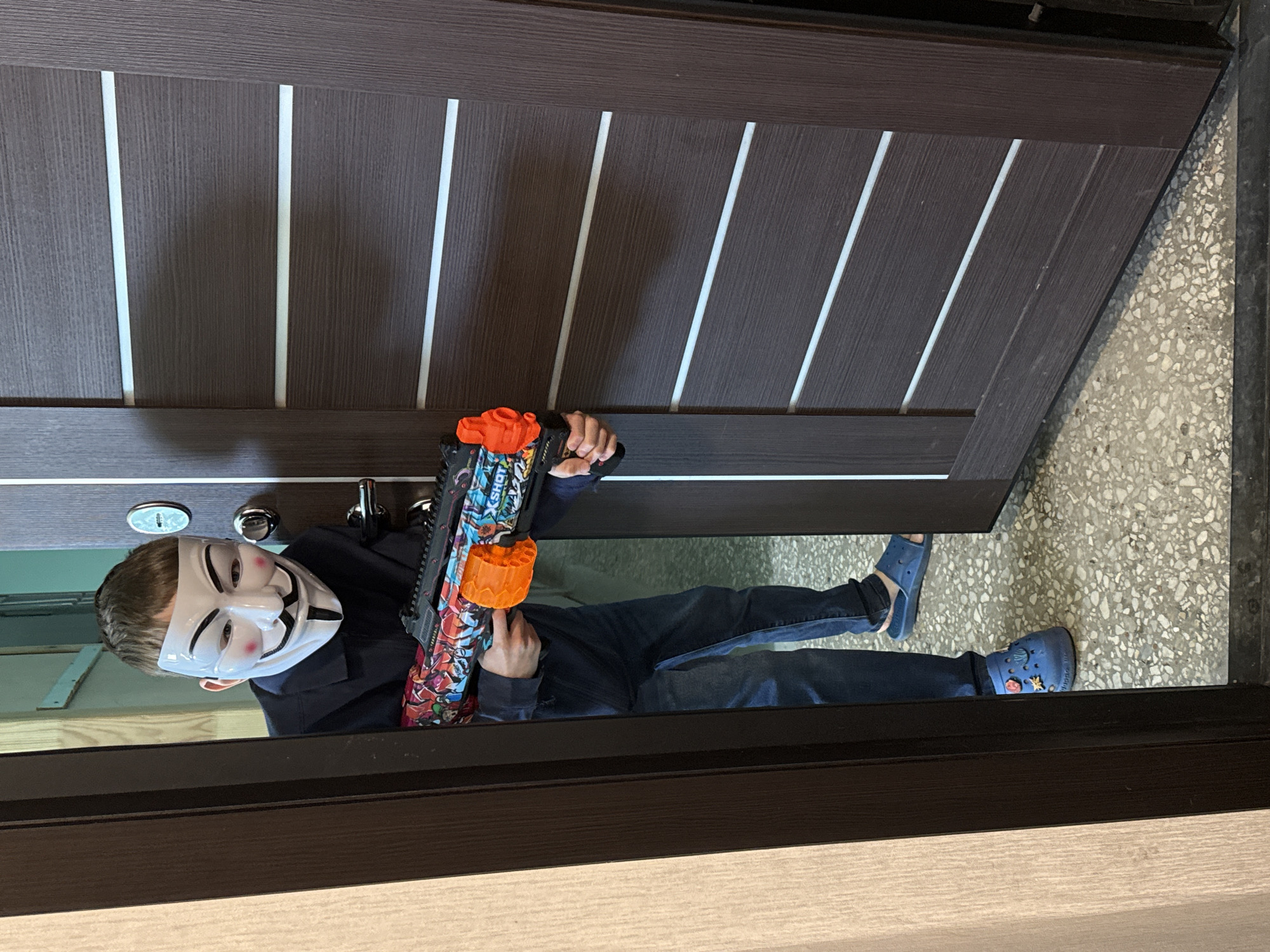 A young person wearing a white Guy Fawkes mask and dark clothing stands in a doorway, holding a colorful toy gun. The person is also wearing blue Crocs with embellishments. The setting appears to be indoors, possibly a home, with a dark brown wooden door and a speckled floor visible. The lighting suggests it is daytime. The person's posture and the mask create a playful or slightly mischievous mood. The toy gun has an orange drum magazine and is decorated with various cartoonish images and text. The person is looking towards the camera.