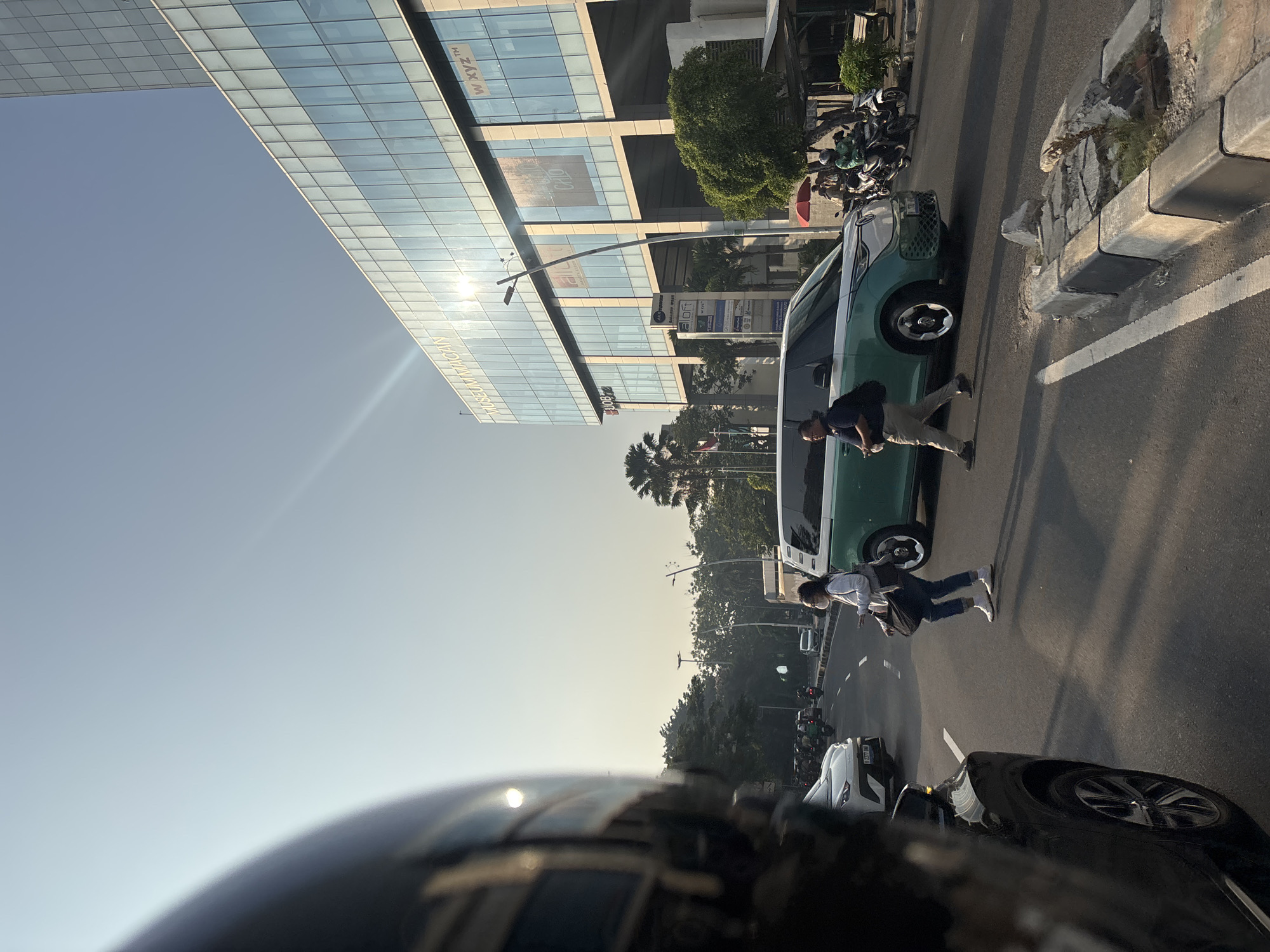 In a street scene in West Jakarta, Indonesia, bathed in the warm glow of a setting sun, a traffic intersection unfolds. A modern, teal-and-white van dominates the foreground, its sleek design contrasting with the surrounding urban environment. To the left of the van, a woman wearing a white shirt, blue jeans, and a mask walks across the street, carrying a dark bag. To the right, a man in a navy shirt and light-colored pants walks in the same direction, also carrying a dark bag. Behind them, a line of motorcycles and cars are visible, suggesting moderate traffic. The background features a multi-story building with a glass facade, reflecting the sky and sun, and a few palm trees dotting the landscape. The street appears to be paved, with a white lane marker indicating a crosswalk. The overall atmosphere is that of a typical urban evening in a bustling Indonesian city.
