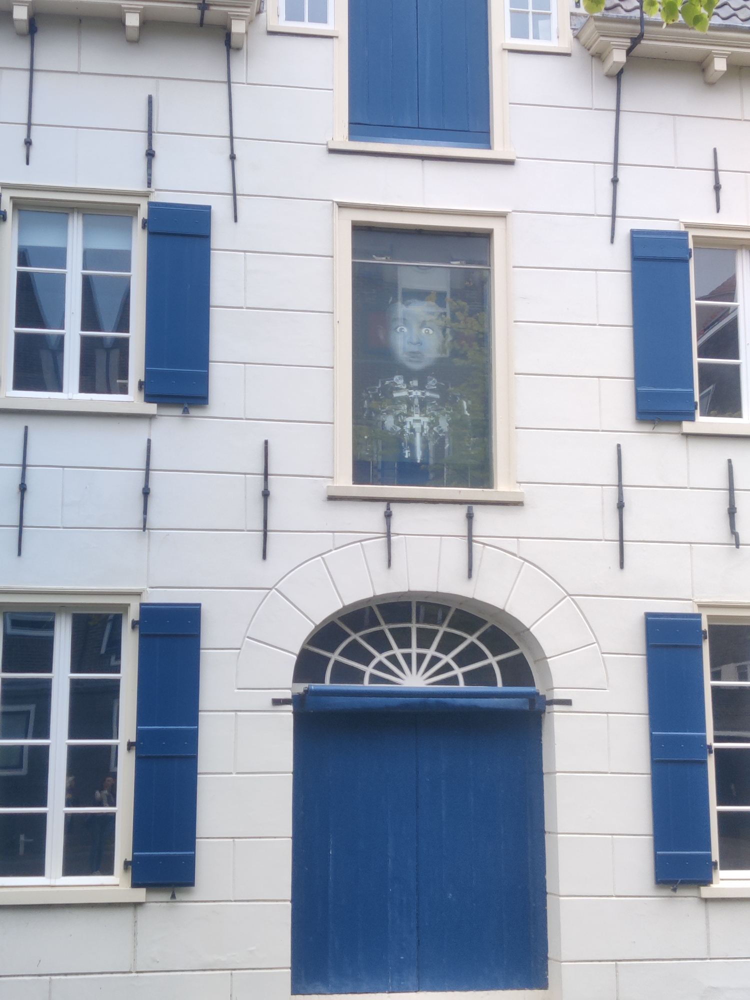 The image shows the facade of a white building with blue shutters and windows. The building is located in the city of Delft, Netherlands. On the second floor, a large window displays a picture of a child's face with wide eyes, superimposed over a robotic structure. Below this window, a large blue door is set within an arched entrance. There are several smaller windows on the first and second floors, all with blue shutters, some of which are closed. The building's facade is made of light-colored stone blocks. The sky is not visible in the frame, but some green foliage can be seen at the top right corner, suggesting it is an outdoor scene. There are no people visible in the image.