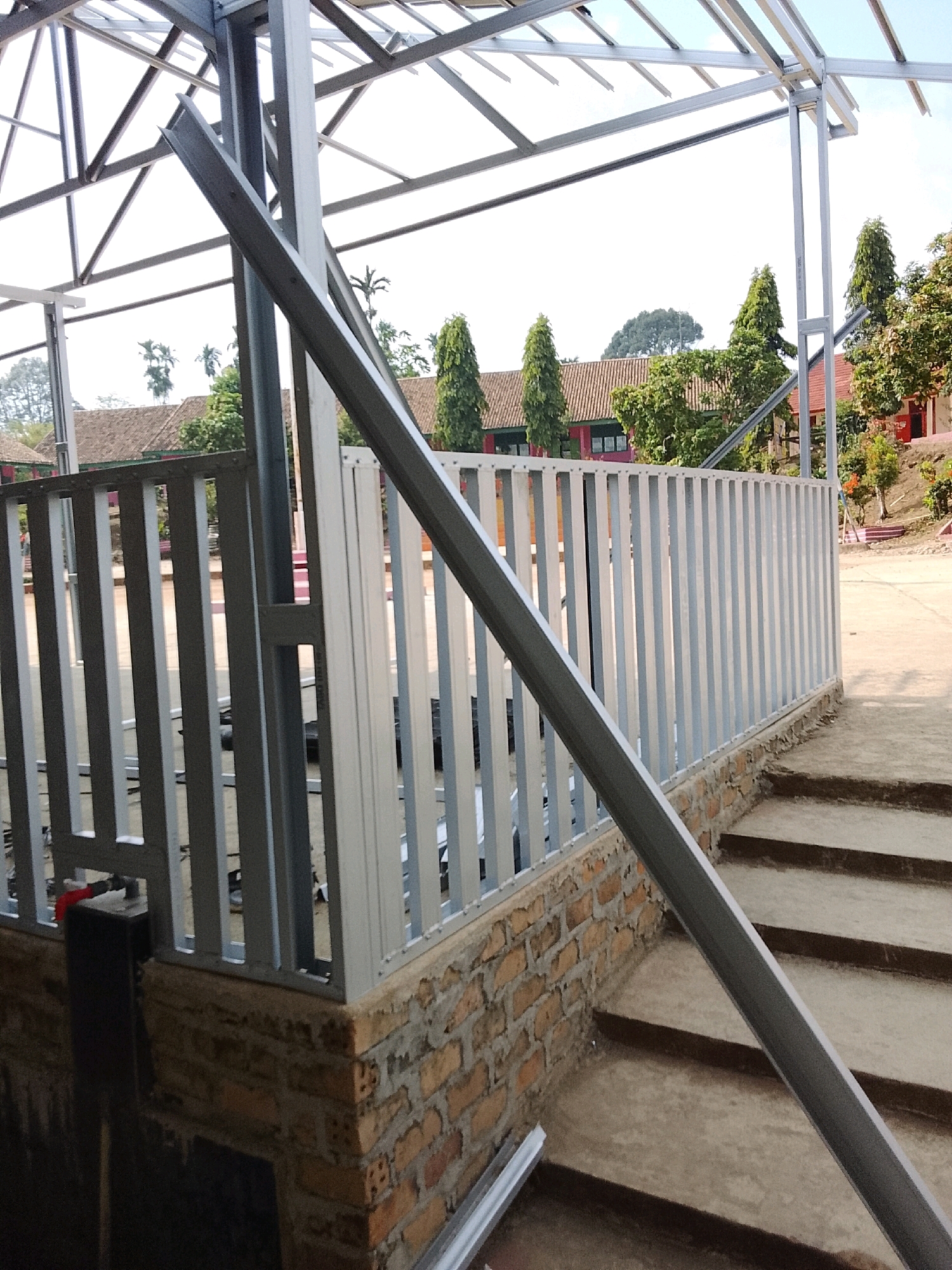 The image depicts the construction of a metal-framed structure, likely a roof or canopy, in what appears to be an outdoor educational facility in Jepara, Indonesia. The foreground features a partially constructed metal fence with vertical slats of alternating gray and white colors, mounted on a low brick wall. Concrete stairs lead up to the level of the fence. A diagonal metal beam, part of the roofing structure, crosses the frame. In the background, several buildings with tiled roofs and some trees are visible. The sky is clear and bright. There are no people or animals present in the image. The scene suggests ongoing construction work at a school or similar public institution.