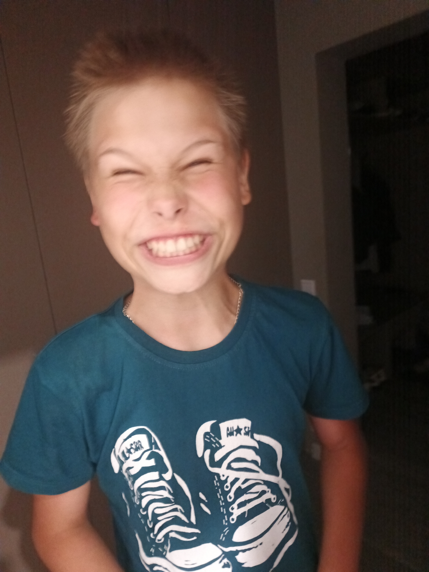 A young boy with light brown, spiky hair is pictured from the chest up, looking directly at the camera with a wide, toothy grin and eyes squeezed shut. He is wearing a teal t-shirt with a graphic print of two white Converse sneakers. A thin silver chain necklace is visible around his neck. The background is dimly lit and appears to be indoors, with a doorway or closet visible on the right side of the frame. There are no specific identifiable landmarks or elements that would indicate a location within Markova, Russia.