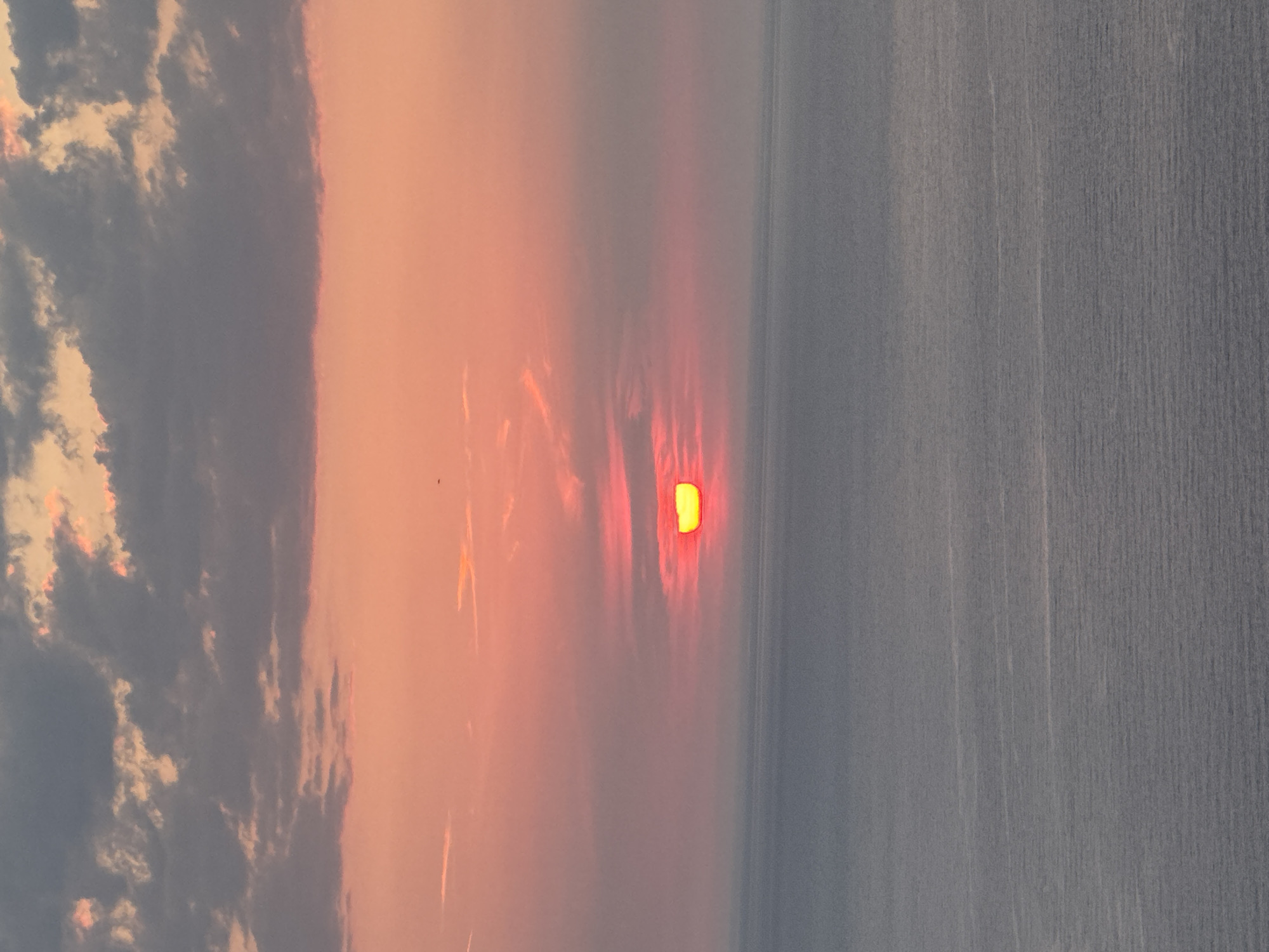 This image captures a serene sunset over the Adriatic Sea, viewed from the vicinity of Zagora, Montenegro. The sky is a gradient of soft pinks and oranges, transitioning into dusky gray clouds. The sun, a bright crescent of yellow and red, is partially submerged on the horizon, casting a warm glow. The sea below is a calm expanse of deep blue, with subtle ripples reflecting the fading light. There are no people, buildings, or explicit signs of human activity visible, emphasizing the natural beauty of the coastal landscape. The scene evokes a sense of tranquility and natural wonder.