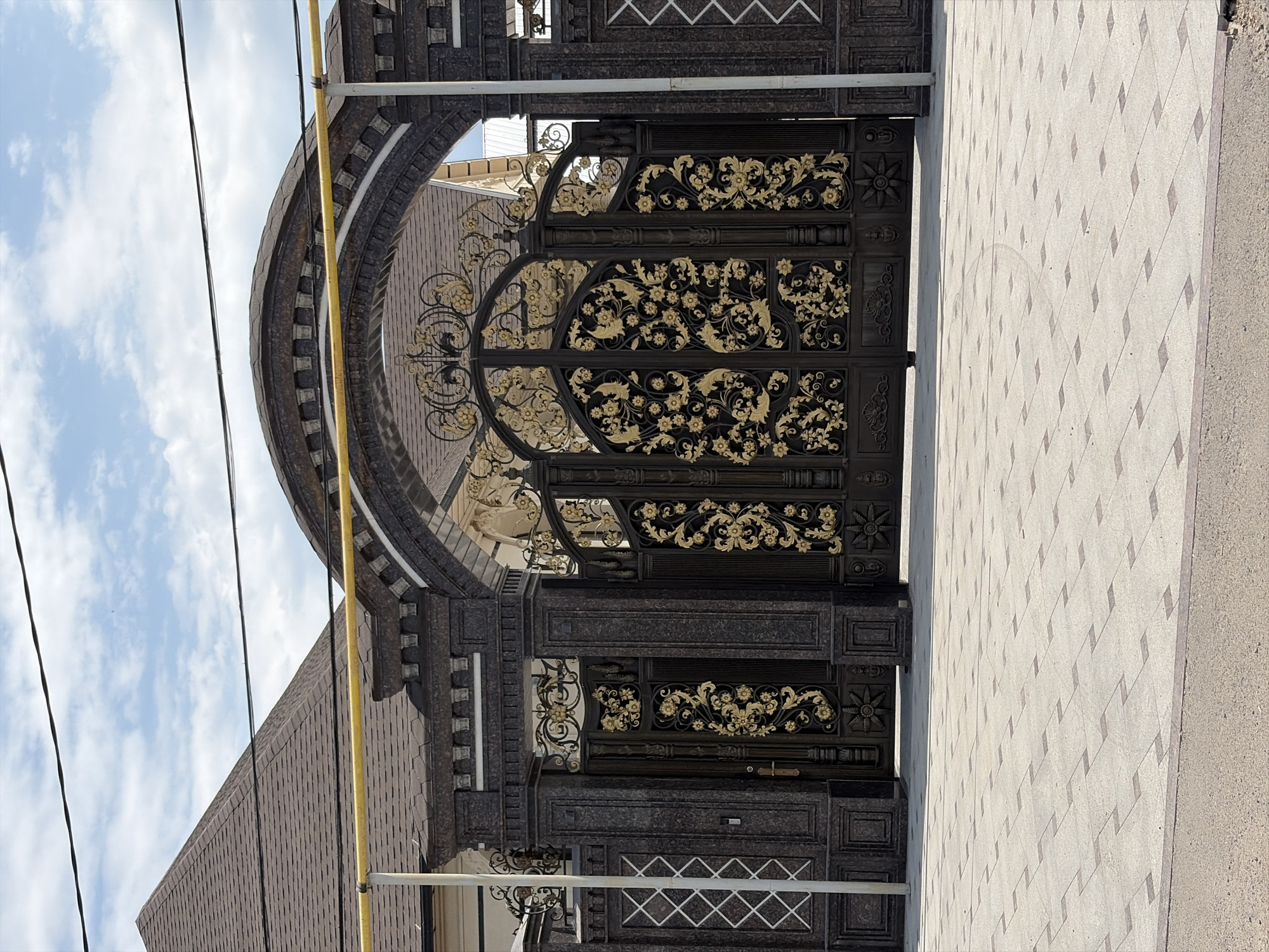 An ornate, dark metal gate with gold floral embellishments is the central focus of the image, situated in front of a building with a dark, textured stone facade and a peaked roof. The gate is arched and appears to be part of a larger fence or wall. The ground in front of the gate is paved with light-colored square tiles arranged in a diamond pattern, with darker accents. A yellow pipe runs horizontally across the top of the scene, supported by posts that blend with the gate's architecture. Overhead, a cloudy blue sky is visible. The scene appears to be an entrance to a private property, possibly a residence or an establishment, in Novopokrovka, Kyrgyzstan. There are no people or animals visible in the image.